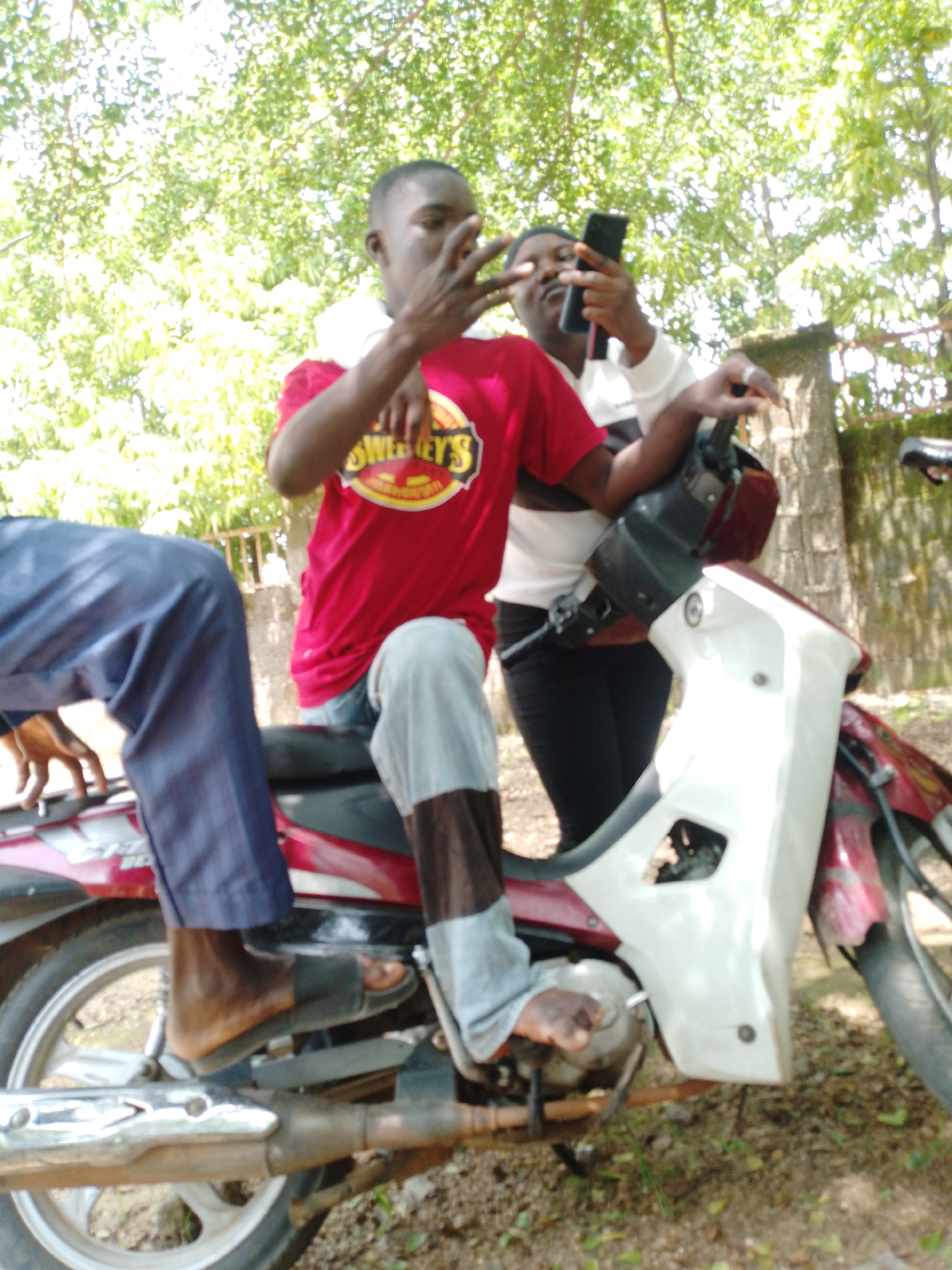In Akwanga, Nigeria, two individuals are pictured near a motorcycle outdoors. The person in the foreground, facing right, is wearing a red t-shirt with "SWEETEY'S" visible on it and gray pants. They are seated on the motorcycle, with their left foot wearing a black sandal resting on the ground. Their right leg is bent, with their foot also on the ground and partially obscured by their pants. The person behind them, facing left, is wearing a white shirt with black accents and black pants. They are holding a black smartphone in their right hand and their left hand is near their face. Both individuals appear to be under a tree with dappled sunlight filtering through the leaves. The motorcycle is red and white, with a visible exhaust pipe and wheel. The ground is covered in dirt and sparse vegetation.