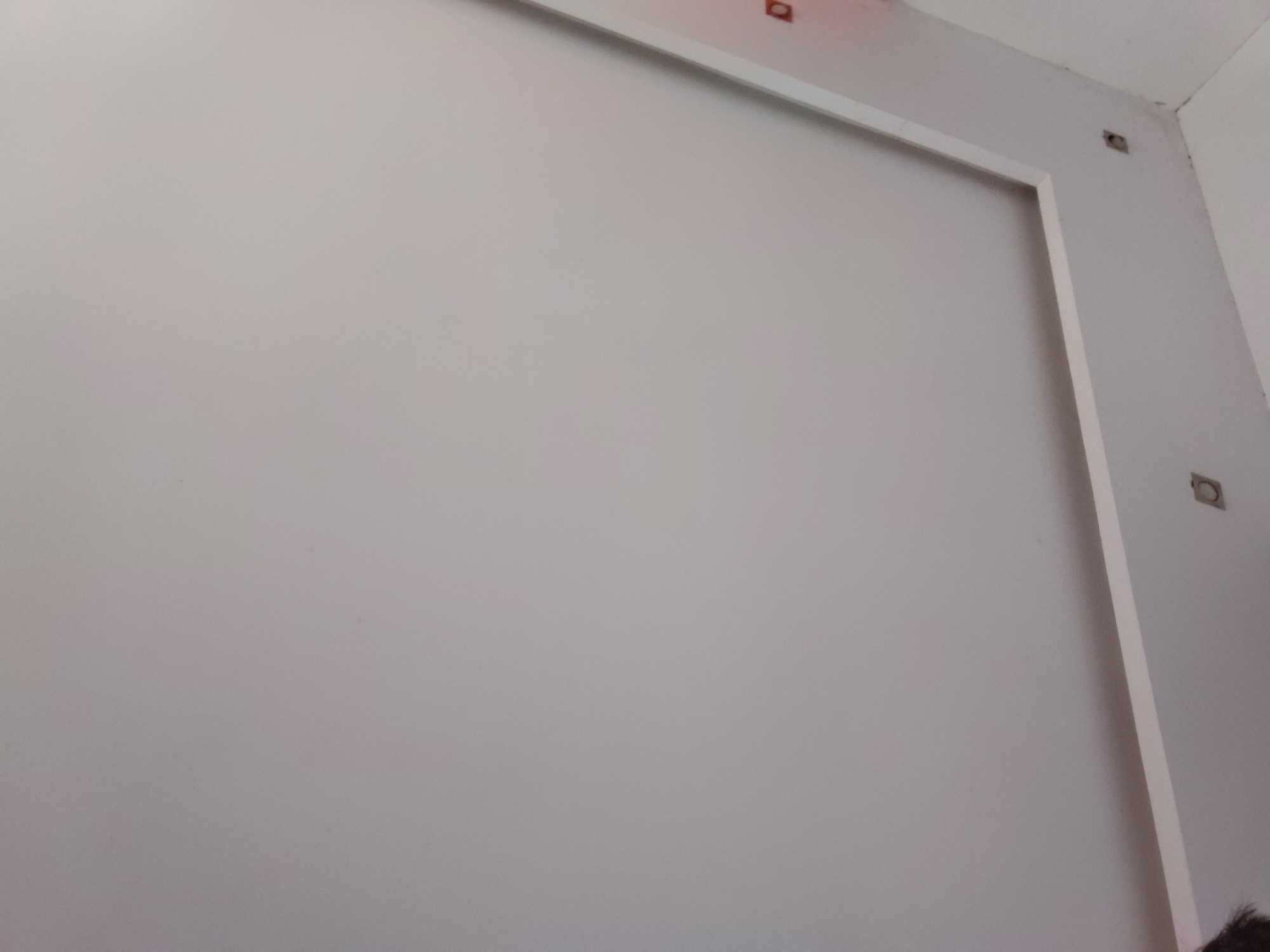 This image captures an indoor scene, likely a room in Santa Ana, Venezuela, featuring a large, light gray wall. A white, rectangular molding or frame is attached to the wall, running horizontally near the top and vertically down the right side, creating a bordered section. The ceiling, which appears white, meets the wall in the upper portion of the frame.
Several square or rectangular cutouts are visible on the upper wall and ceiling. These appear to be empty electrical boxes or mounting points for fixtures, suggesting the room is either under construction, undergoing renovation, or awaiting the installation of lighting, speakers, or other utilities. Above the horizontal white frame, on the ceiling, there are also some reddish-brown marks, possibly related to construction or priming.
A small portion of dark hair is visible at the very bottom right corner of the original image, indicating the presence of a person, likely the photographer. The overall ambiance is clean and understated, with a dominant color palette of light gray and white. No specific activity is depicted, but the exposed electrical elements suggest a work-in-progress or an unfurnished space. There is no visible text in the image.