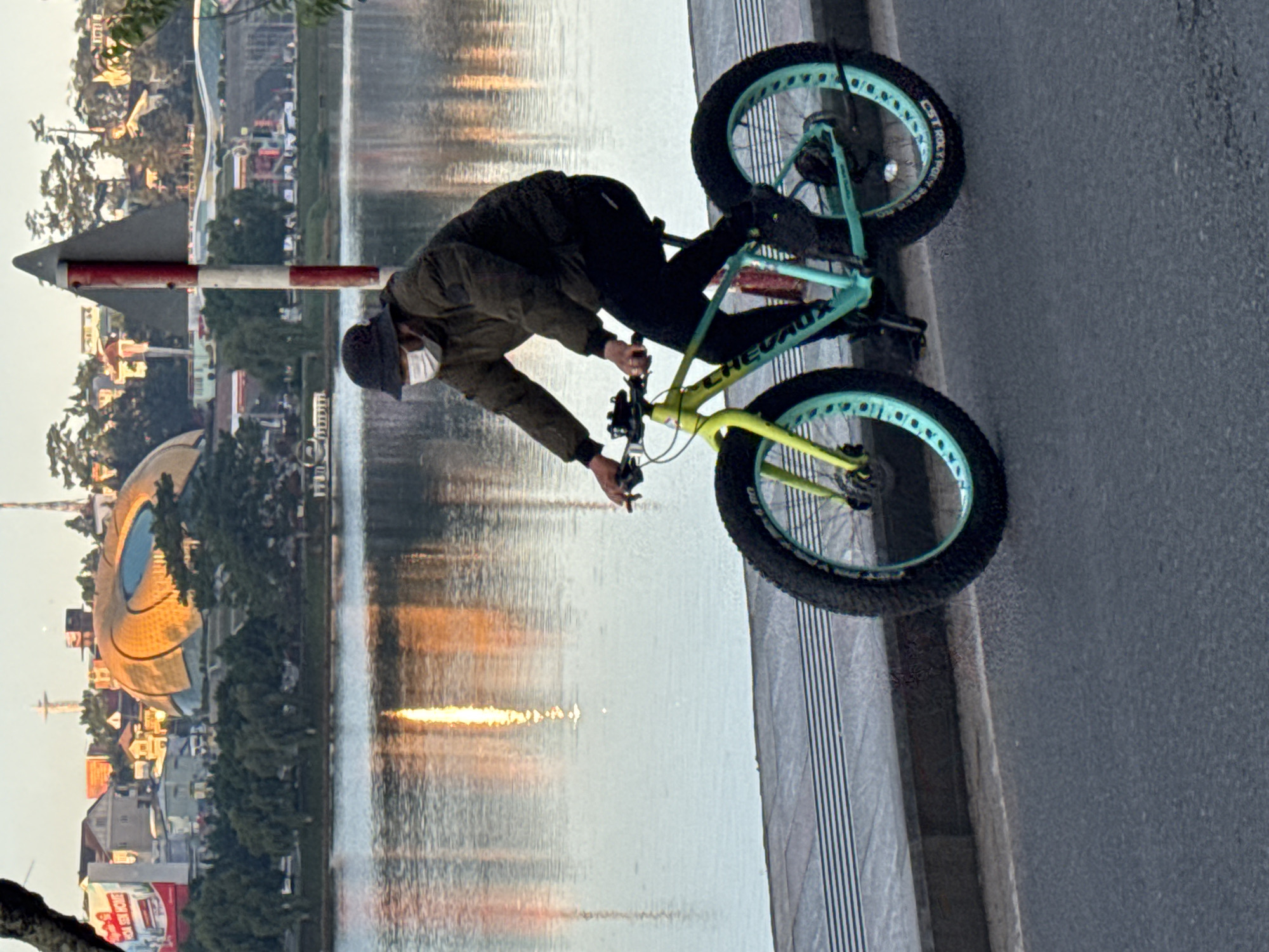 A person wearing a dark hat and a white face mask rides a fat-tire bicycle with a neon yellow frame and aqua rims on a paved path adjacent to a body of water. The cyclist is wearing a dark jacket and dark pants. In the background, across the water, is a cityscape with buildings, trees, and a distinctive golden, dome-shaped structure. The water reflects the sunlight and the buildings. A red and white striped pole stands to the right of the cyclist. The scene appears to be set in the Phường Xuân Hương area of Đà Lạt, Vietnam, known for its Xuan Huong Lake, which is likely the body of water depicted.
