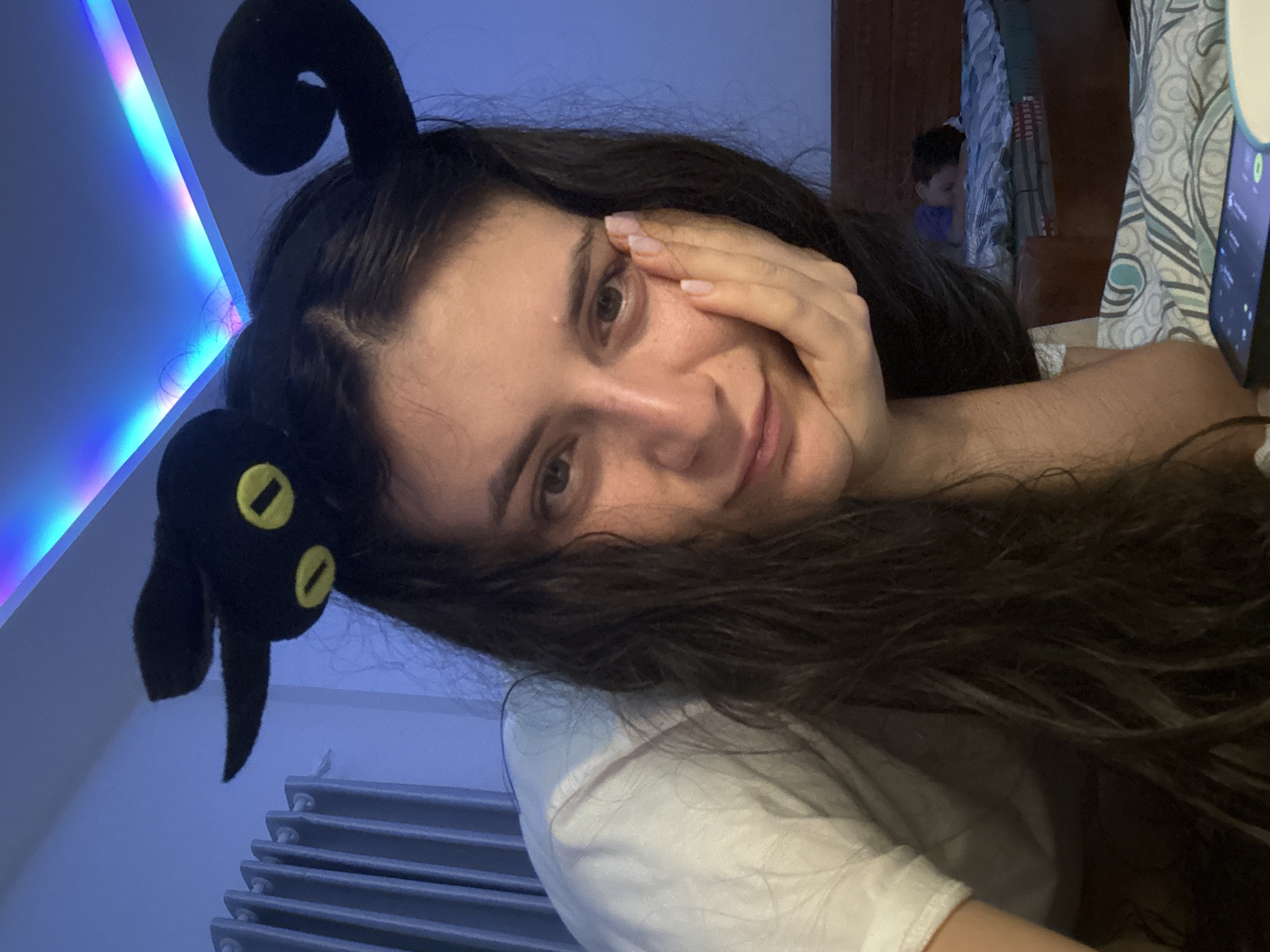 A woman is posing for a selfie or photo. She has long, dark hair and is wearing a black headband with black plush objects resembling insect antennae or horns. She is holding her hand to her face.
Behind her is a room with light blue walls and a radiator. A child can be seen in the background, seated in a room with wooden furniture and a patterned fabric. To the right, a mobile phone is visible in the foreground.
There is no information to suggest that this image was taken in the city of Mũi Né, Vietnam.