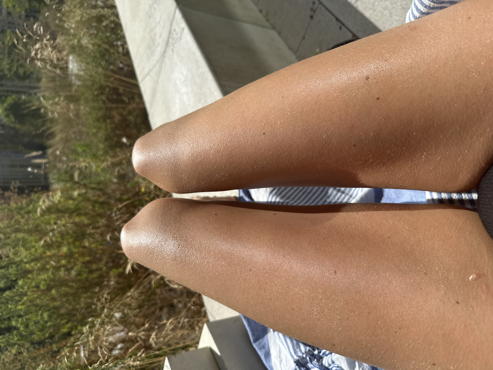 A close-up, ground-level view focuses on two tanned, sun-kissed legs positioned on a blue and white striped towel. The legs are illuminated by bright sunlight, highlighting a subtle sheen on the skin, suggesting recent exposure to water or lotion. The background features dry, golden grasses and foliage, with hints of concrete structures and a sunlit paved area. The scene evokes a sense of relaxation and outdoor leisure in a warm climate. There is no discernible information within the image to indicate the location as Amsterdam, Netherlands.
