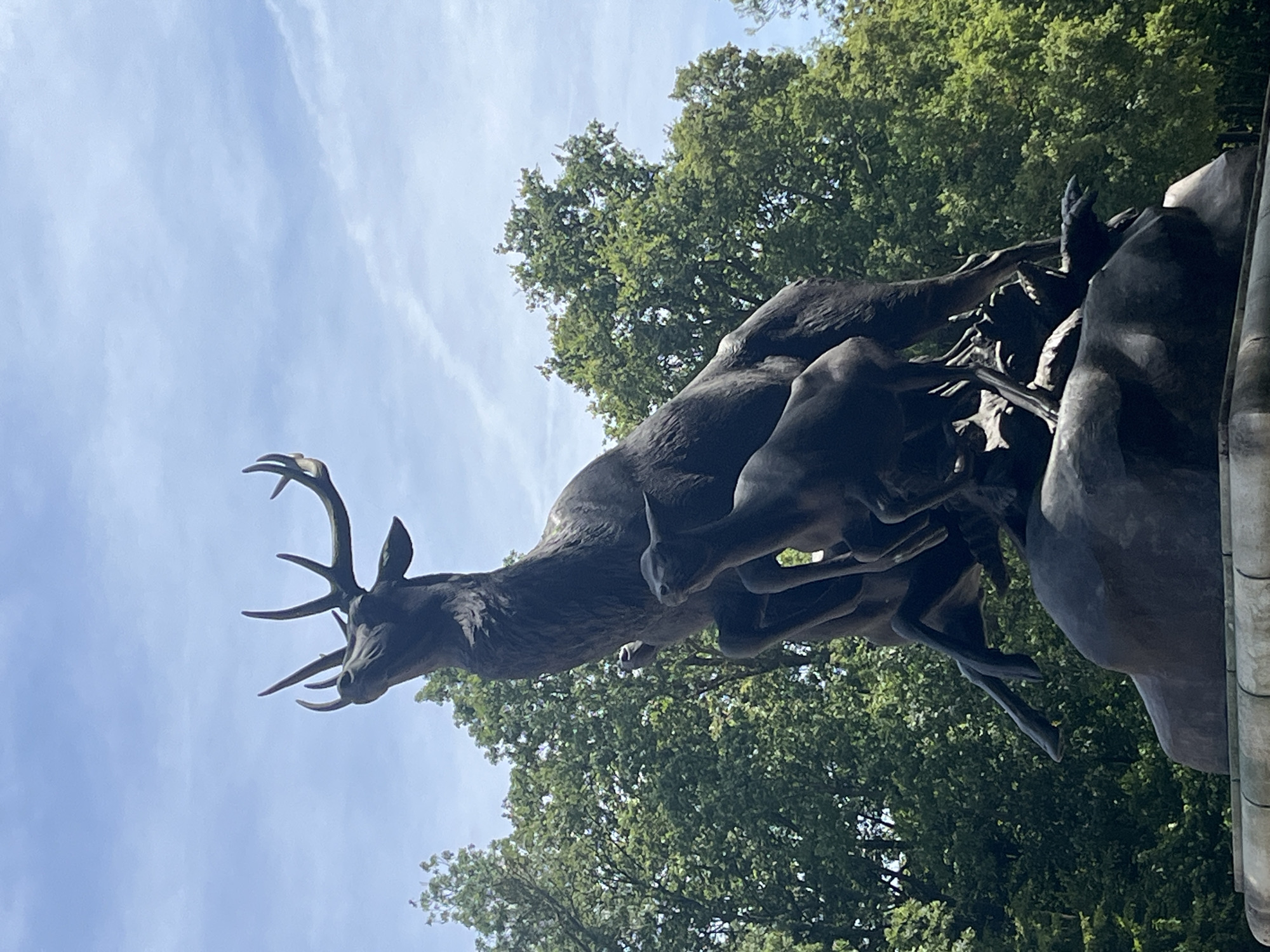 The photograph depicts a bronze sculpture of a stag with prominent antlers, rearing upwards. Below the stag, two smaller deer are positioned, one seemingly sheltering beneath the stag. The sculpture rests on a stone base, which is partially visible at the bottom of the frame. Lush green trees form the background, and a bright blue sky with scattered white clouds is visible above. This is the "La Harde" monument located in Parc de Sceaux, Antony, France.
