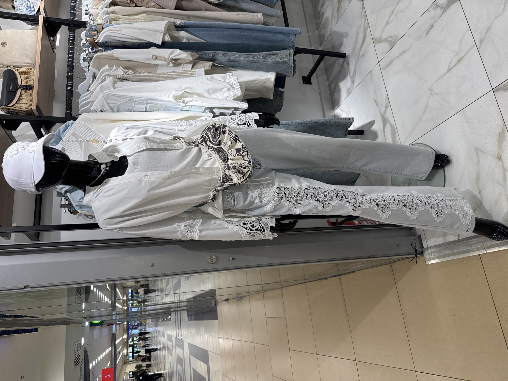 A clothing display in a shop, potentially in Saint Petersburg, Russia, showcases a mannequin wearing light blue jeans with white lace trim on the cuffs, paired with a white outfit and a white head covering. The mannequin stands behind a glass panel, reflecting an indoor shopping area with lights and people. Racks of clothing are visible in the background of the clothing display. The scene's setting appears to be a commercial environment like a shopping mall.