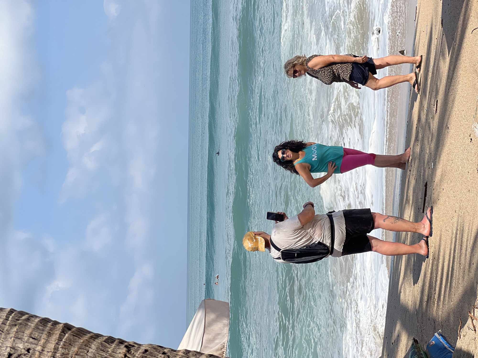 In Kuta, Indonesia, a man wearing a yellow cap, a beige t-shirt, black shorts, and a backpack, stands on a sandy beach holding a smartphone. He is positioned facing the ocean and appears to be taking a photograph. To his right, two women are standing by the water's edge. The woman on the left wears a teal tank top with the word "MOO" printed on it in white, and pink capri leggings. The woman on the right is dressed in a patterned sleeveless top and dark shorts. The ocean exhibits waves, and the sky is partly cloudy. A large tree trunk is visible on the left side of the frame, casting shadows on the sand.