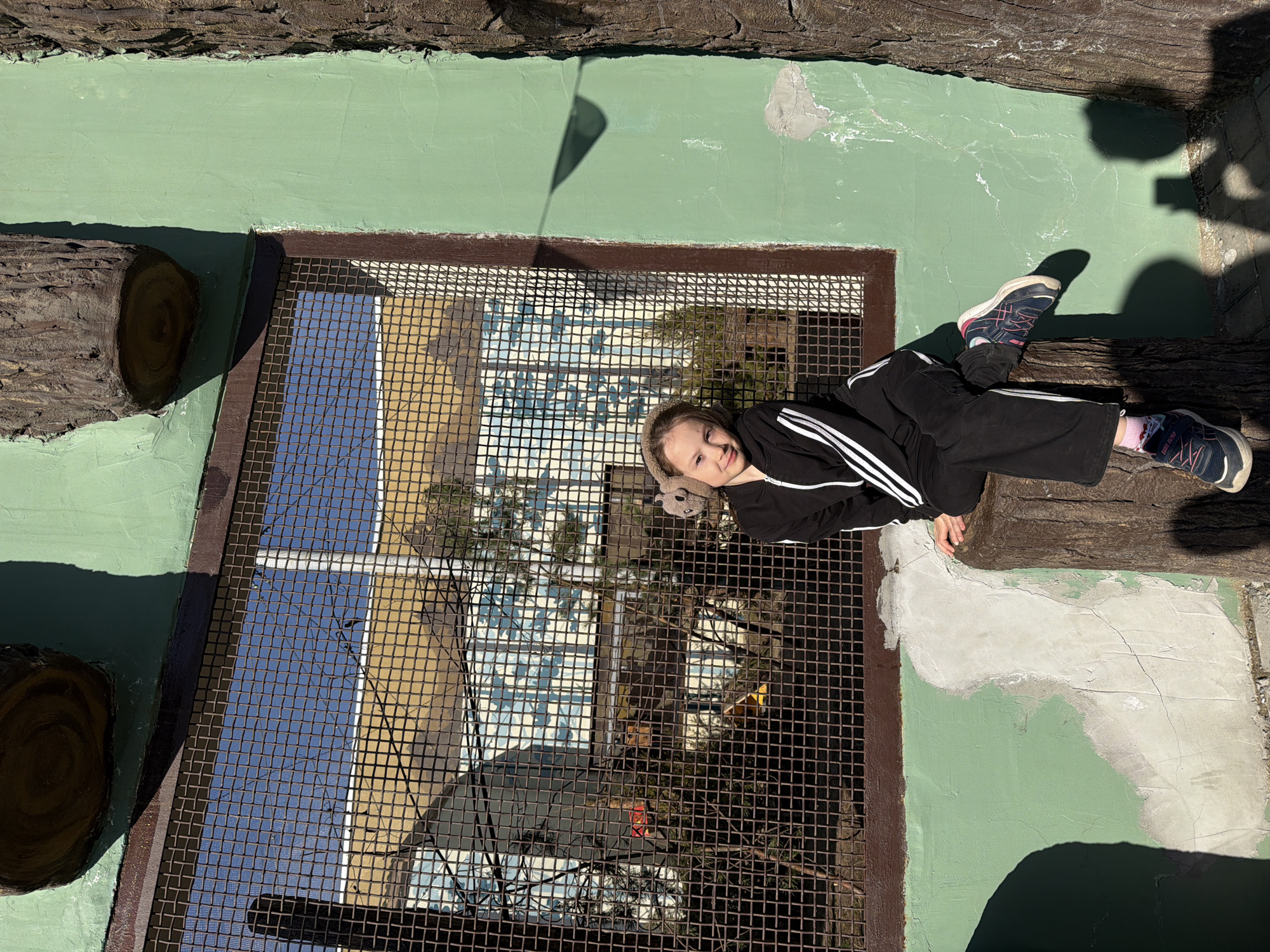 The image shows a girl lying on a metal grate. She is wearing a black tracksuit with white stripes and athletic shoes. Her head rests near the center of the grate, and her legs extend towards the top right corner of the frame.
Below the grate, there is a reflection of buildings and trees.
The area surrounding the grate is painted green and is bordered by what appear to be artificial logs.
The image was taken at the Perm Zoo in Perm, Russia.