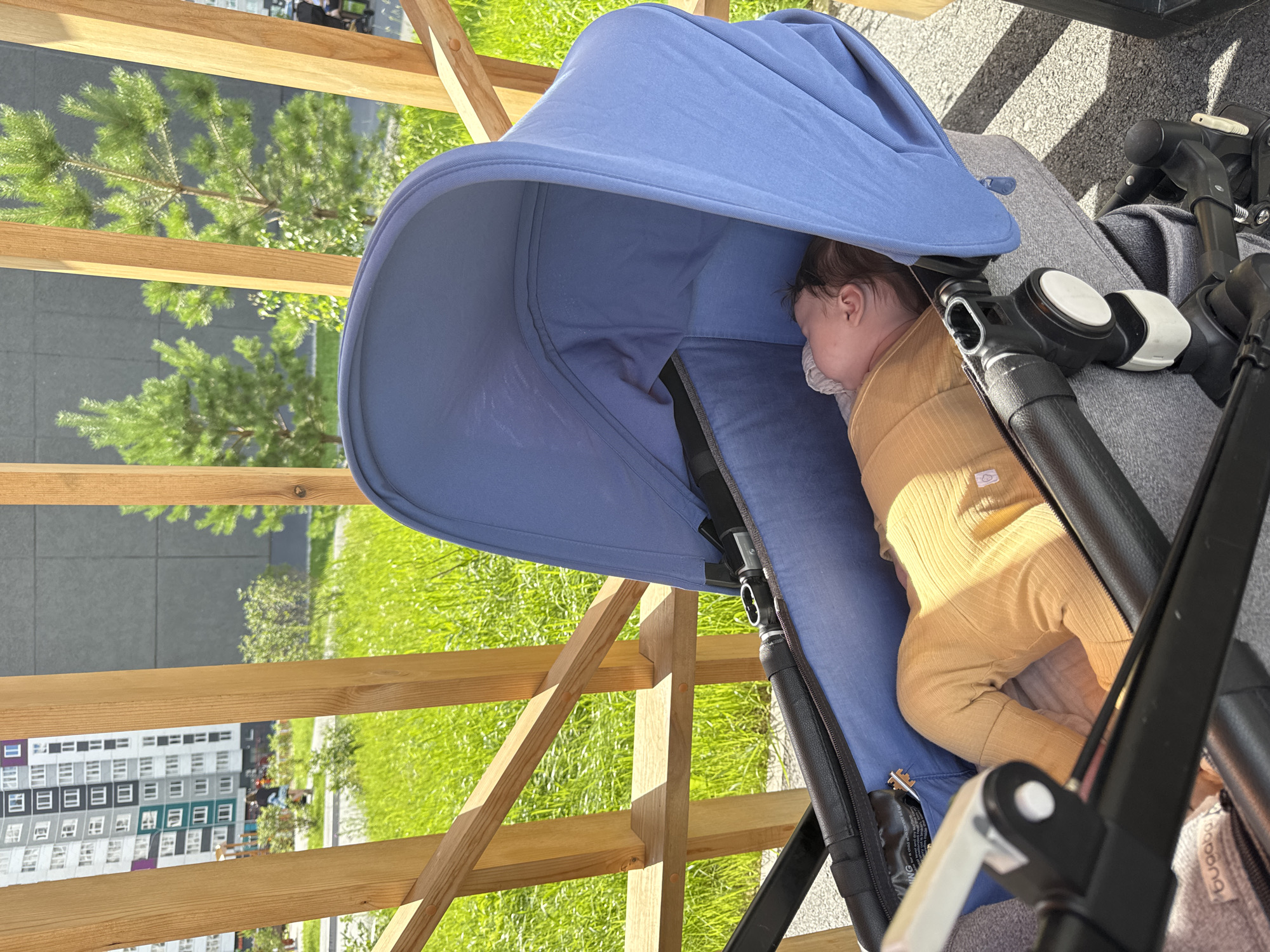 The image shows a baby sleeping in a blue stroller, outdoors. The stroller is partially covered by its hood, and the baby is wearing a tan ribbed onesie. The background features a wooden fence, green grass, and a building with many windows. Sunlight casts shadows on the ground, suggesting it's daytime. The scene appears peaceful, with the baby comfortably asleep. The location is identified as Patrusheva, Russia. There is no visible text in the image.