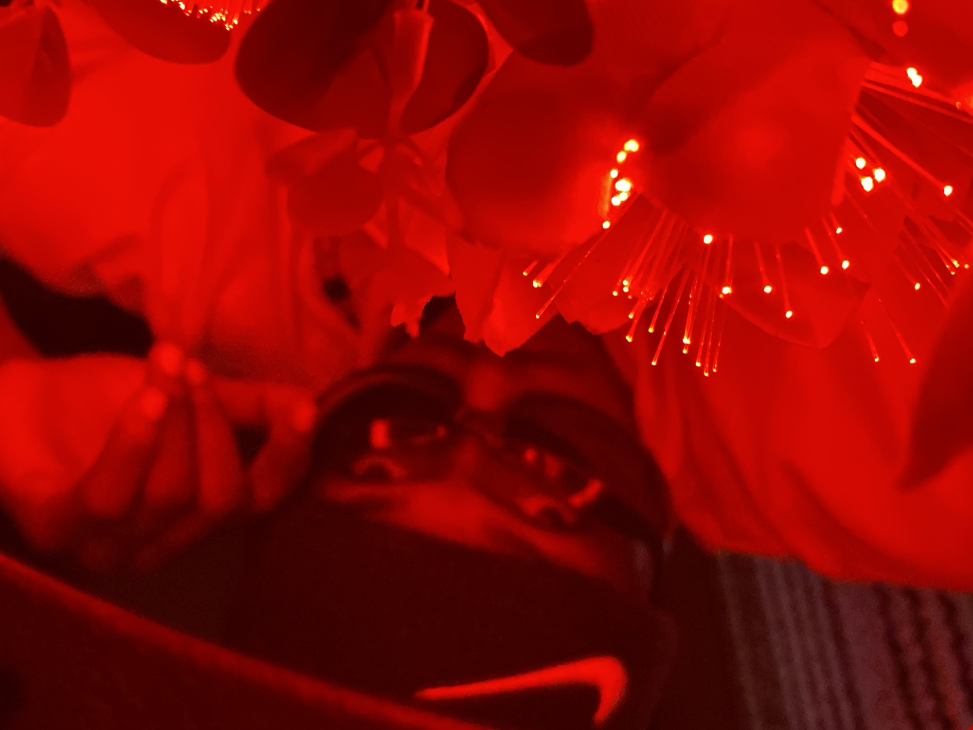 The image shows a close-up view of a person wearing a black cap with a red Nike logo and dark sunglasses. The person is illuminated by red light, which also casts a strong red hue over the surrounding objects, which appear to be floral or decorative with small, glowing fiber optic-like strands. The background is dark and blurred, with some vertical lines suggesting a textured surface. The overall impression is one of a stylized, dimly lit indoor setting. There are no clear indicators of the specific location within Abakaliki, Nigeria.