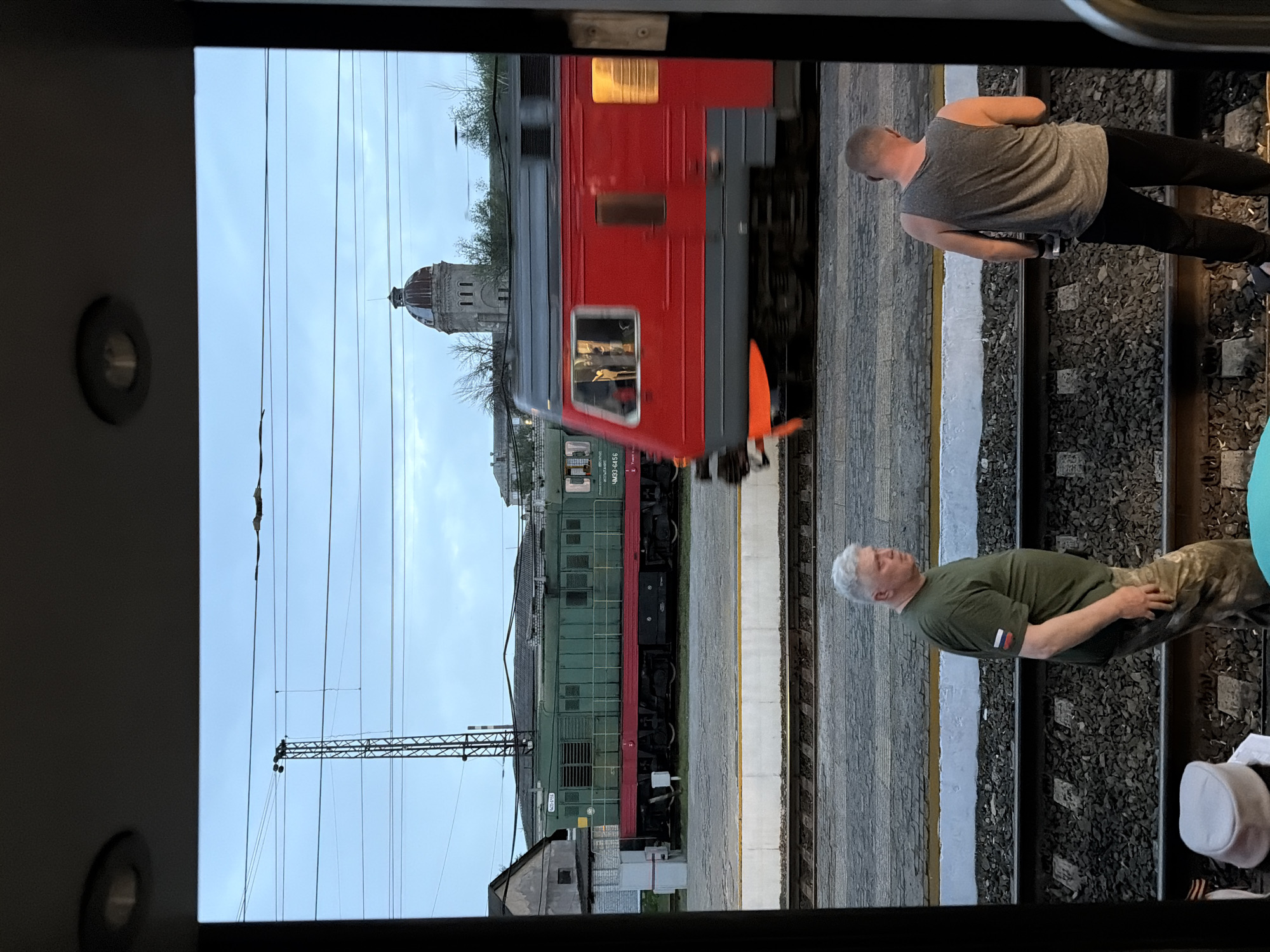 The image, likely taken from a train window, shows a station platform with train cars passing. A building with a prominent dome is visible in the background, indicating the location as Rtishchevo, Russia. Two men are on the platform. One man is wearing a tank top and dark pants, the other is wearing a green t-shirt and camouflage pants. The man in the green shirt has a Russian flag emblem on his sleeve.