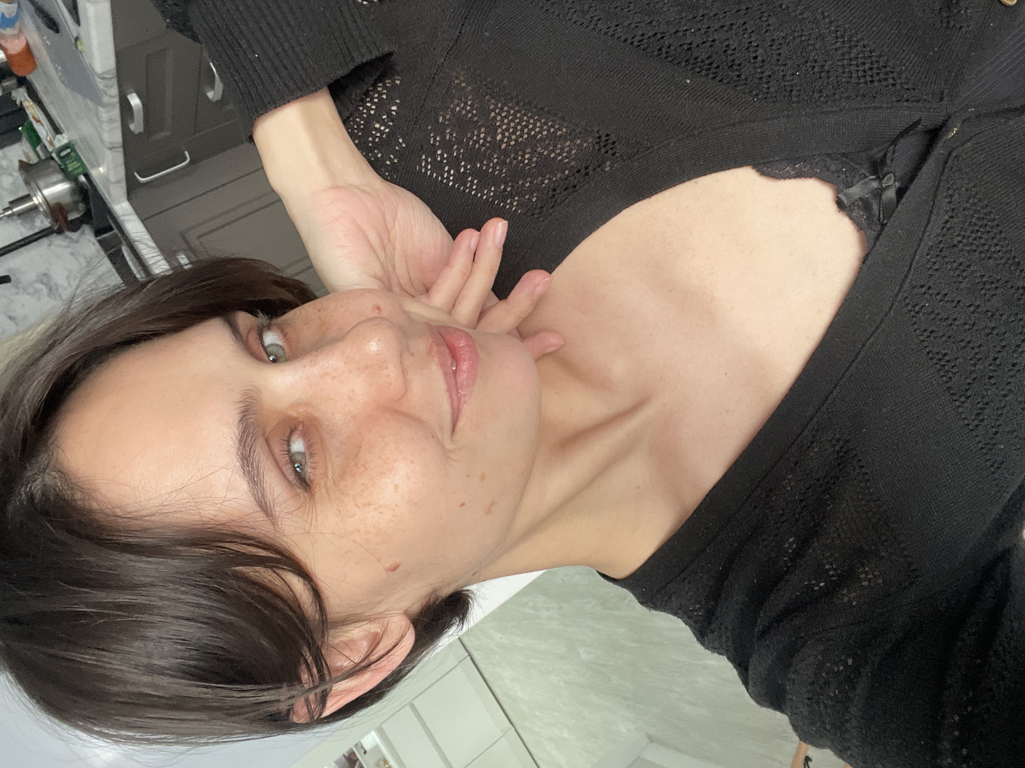A young woman with short, dark brown hair and fair skin with freckles poses for a selfie-style photograph. Her eyes appear light-colored. She is wearing a black, open-front knit cardigan or top. Her left hand is raised and positioned near her face. In the background, parts of what appears to be a kitchen are visible, including countertops, cabinets, and stainless steel kitchenware. The environment suggests an indoor setting, likely a domestic residence. There is no evidence to support the location in Izhevsk, Russia.