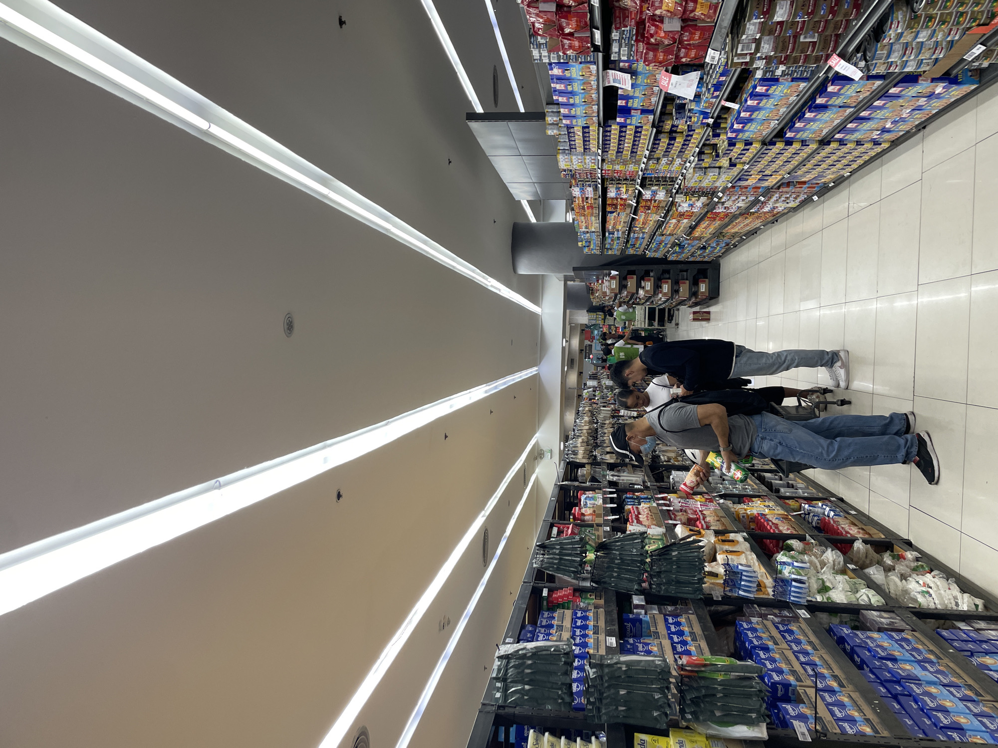 The image depicts an interior view of a supermarket aisle in Davao City, Philippines. The aisle is well-lit by long, linear fluorescent lights on the ceiling. Shelves on both sides of the aisle are stocked with various packaged goods, including canned foods, boxes of cereal or other dry goods, and what appear to be bags of snacks or coffee. The flooring is tiled in a light color.
In the foreground, a man wearing a black baseball cap, a grey t-shirt, blue jeans, and black athletic shoes is standing and looking at items on a shelf. He is wearing a light blue disposable face mask. Next to him and slightly behind, a woman with dark hair, wearing a white t-shirt, is visible. Further down the aisle, another man wearing a black jacket and dark pants is looking at his phone. Several other shoppers and store employees are visible further down the aisle, some pushing shopping carts. One individual in a green shirt appears to be an employee. The overall atmosphere suggests a typical grocery shopping experience.