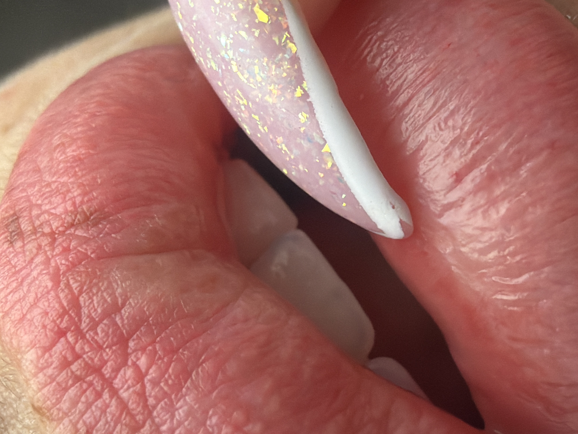This close-up photograph captures an intimate detail of a person's mouth interacting with a manicured fingernail. The image primarily features a section of human lips, displaying their natural texture with fine lines and a reddish-pink hue. Between the lips, several white teeth, likely lower front teeth, are visible. A long, oval-shaped fingernail, painted in a light pink base with numerous iridescent glitter flakes (appearing golden or yellow) and a distinct white French tip, is positioned within the mouth, touching the inner lip and possibly the teeth. The white tip of the nail shows a slight textured or raised edge. The background is dark and blurred, drawing full attention to the main subjects. The exact activity is ambiguous but involves the nail making contact with the mouth. No specific emotions are discernible, and no text is visible. While the image itself lacks explicit location cues, the context provided indicates it is set in Saint Petersburg, Russia.