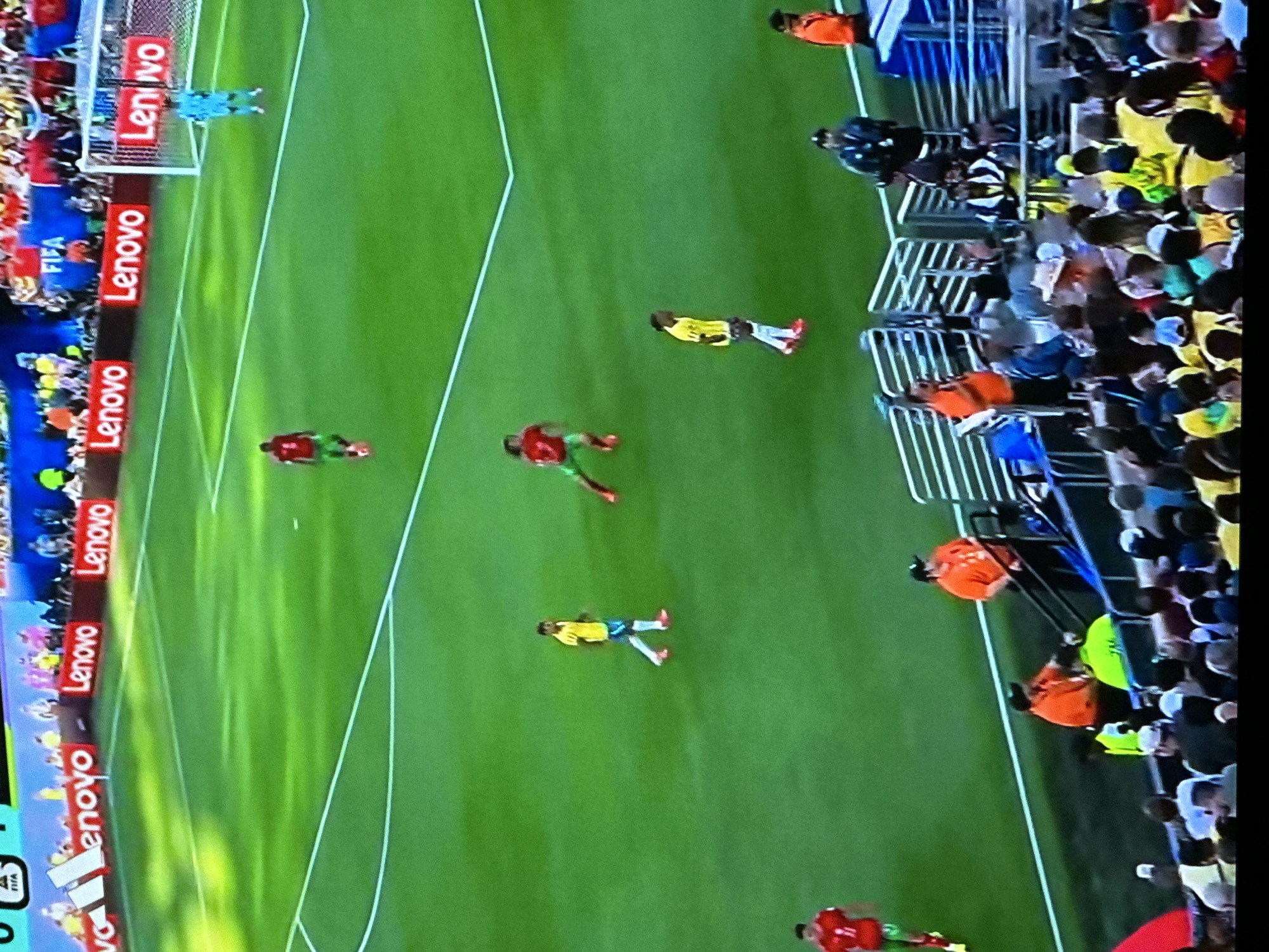 The image depicts a soccer match in progress on a vibrant green field. A group of players are visible, some in red and green uniforms, others in yellow and blue. One player in a red jersey with green shorts runs towards the left of the frame, while another in a yellow jersey with blue shorts stands near the center. In the background, a goalkeeper in a light blue uniform is positioned near the goal. Spectators are seated in the stands behind a barrier. The field is marked with white lines, and advertisements for "Lenovo" are visible along the perimeter. The overall scene suggests an active sporting event. The mention of Gura Top, Nigeria, is an external detail not inferable from the image itself.