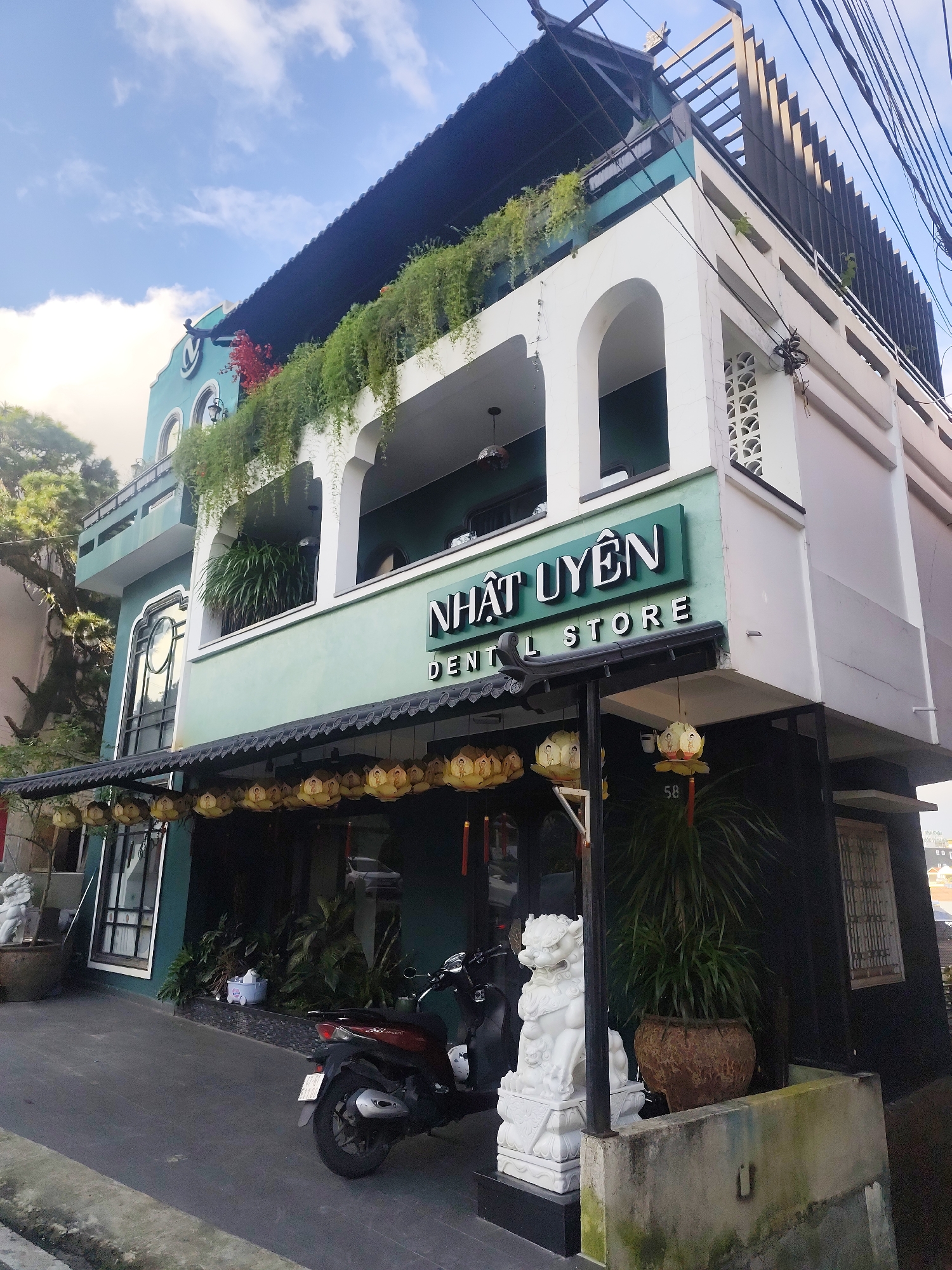 The image depicts the exterior of a two-story building with a green facade, located in Da Lat, Vietnam. The building houses a dental store. The facade features arched windows and balconies adorned with lush green hanging plants. A prominent sign with white lettering reads "NHAT UYEN DENTAL STORE". Decorative lanterns in a lotus flower design hang from the overhang of the building. In the foreground, a dark-colored scooter is parked on the paved ground. Two white lion statues stand guard near the entrance. The sky above is blue with scattered clouds. The overall impression is of a well-maintained establishment with a touch of traditional Vietnamese architectural elements.