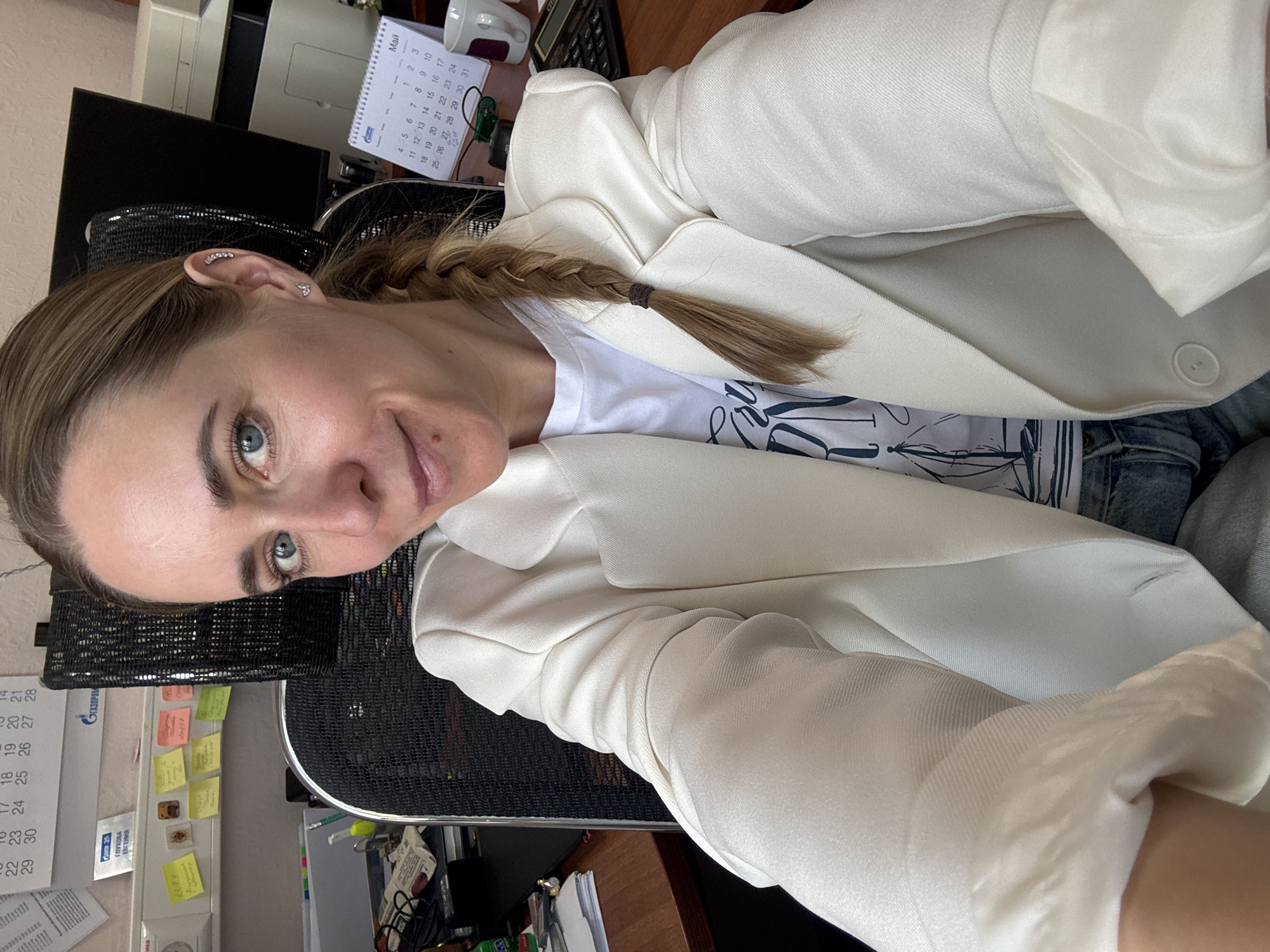 Here is a detailed description of the image:
**Scene:** The image appears to be a selfie taken by a woman at her desk, likely in an office setting. The orientation is slightly tilted.
**People:** A woman with light brown hair pulled back in a braid is the main subject. She has blue eyes and fair skin. She is wearing a white blazer over a white t-shirt.
**Objects:**
* **Desk:** A wooden desk is visible.
* **Office supplies:** A printer, a calendar, a calculator, sticky notes, and other papers are scattered on the desk. The calendar shows the month of May, and one of the papers has the name "ГЛУХОВА ЕВГЕНИЯ" (Glukhova Evgenia) printed on it.
* **Chair:** A black mesh office chair is partially visible.
* **Earrings:** The woman has multiple earrings on her left ear.
**Environment:** The presence of office equipment and supplies indicates the photo was taken in a workplace. Based on the calendar, it is likely May 2023.
**Location:** Chaikovsky, Russia can be assumed due to the Russian text and the presence of Газпром information visible in the photo, indicating a Gazprom-related office.
**Actions/Interactions:** The woman is holding the camera at arm's length and looking at the lens, suggesting she is taking a selfie.