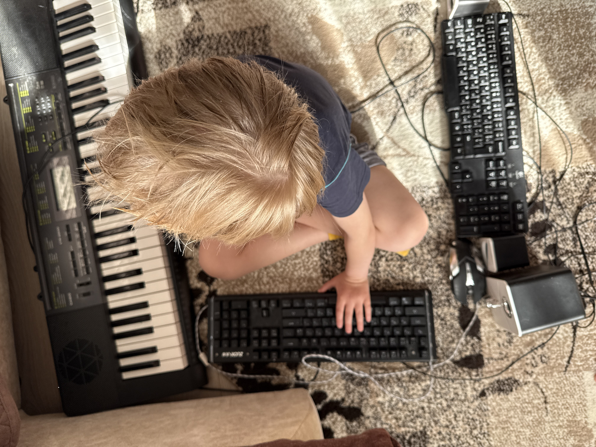 An overhead photograph, taken in Марковский, Russia, depicts a young child seated on a patterned rug, surrounded by various electronic devices. The child, with light blonde hair, is wearing a dark blue t-shirt with a light blue stripe on the sleeve and light-colored shorts. Their right hand rests on a black full-size computer keyboard, branded "DURANDAL," positioned in front of them. To the child's immediate left is a black electronic musical keyboard with white and black keys, identified as a "CASIO" brand instrument. Further to the child's right and slightly behind them, another black computer keyboard, possibly a "Logitech" model with Russian characters, is situated on the rug. Nearby, a black and silver computer mouse and a silver-topped, black-bodied audio device or speaker are also present. Multiple black and white cables interconnect these electronic components, sprawling across the beige and dark brown patterned rug. Portions of light-colored upholstered furniture are visible on the left and bottom edges of the frame, indicating an indoor domestic setting. Direct sunlight casts visible shadows across the rug.