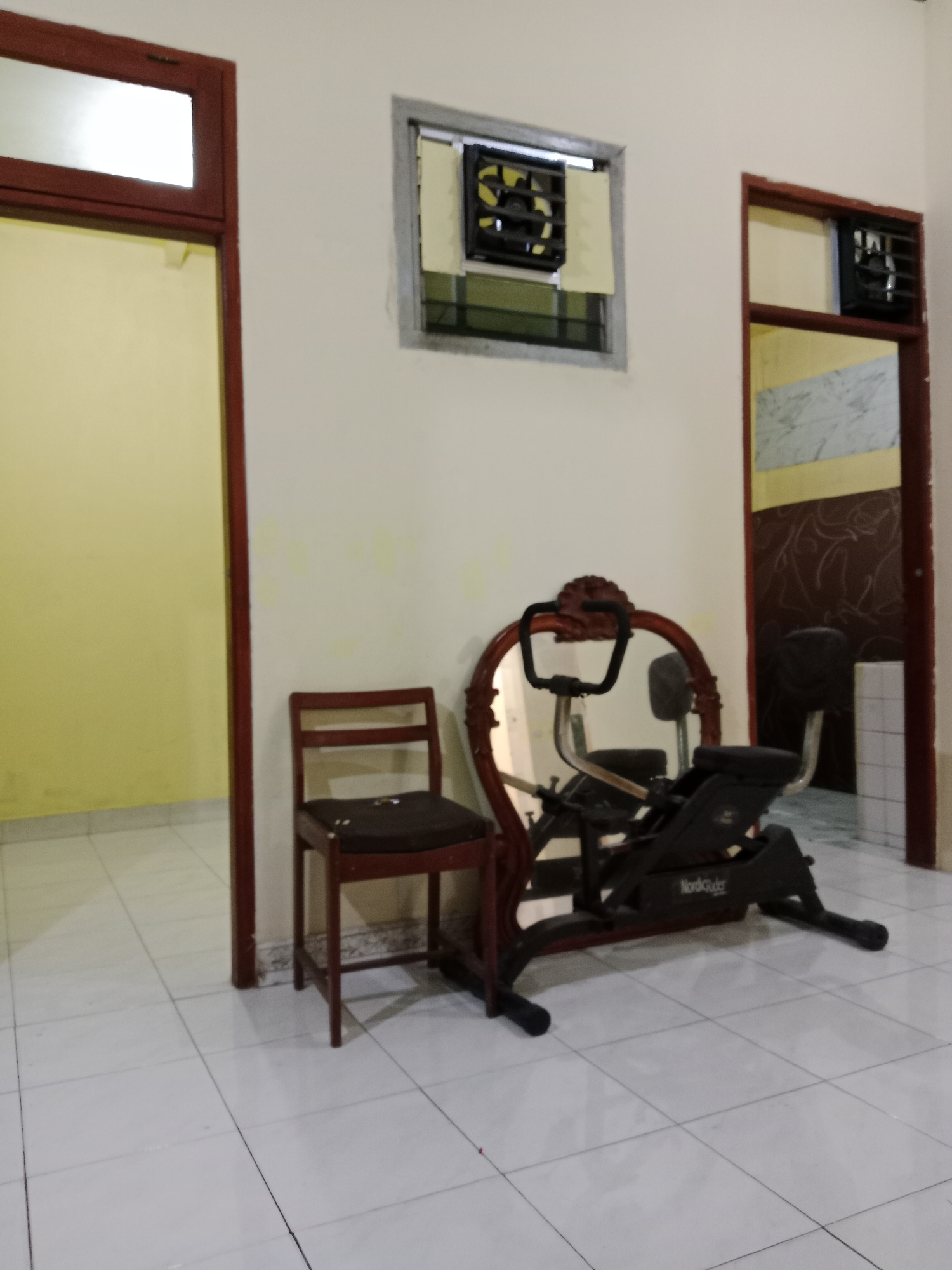 The image depicts an indoor scene, likely a hallway or room with tiled flooring. On the left, a doorway with a reddish-brown frame leads into a room with yellow walls. In the center, a dark-colored wooden chair with a brown cushion stands next to an elliptical exercise machine. The exercise machine is dark gray and has a wooden frame around its mirror. Above the exercise machine, a mirror with an ornate wooden frame is positioned against the wall. To the right of the exercise machine, another doorway with a reddish-brown frame is visible, leading to a room with patterned wallpaper and a ventilation fan in the wall. A second ventilation fan is present in the wall above and to the left of the exercise machine. The overall impression is of a functional space, possibly within a residential or commercial building in West Jakarta, Indonesia, that has been repurposed for exercise or is awaiting further setup.