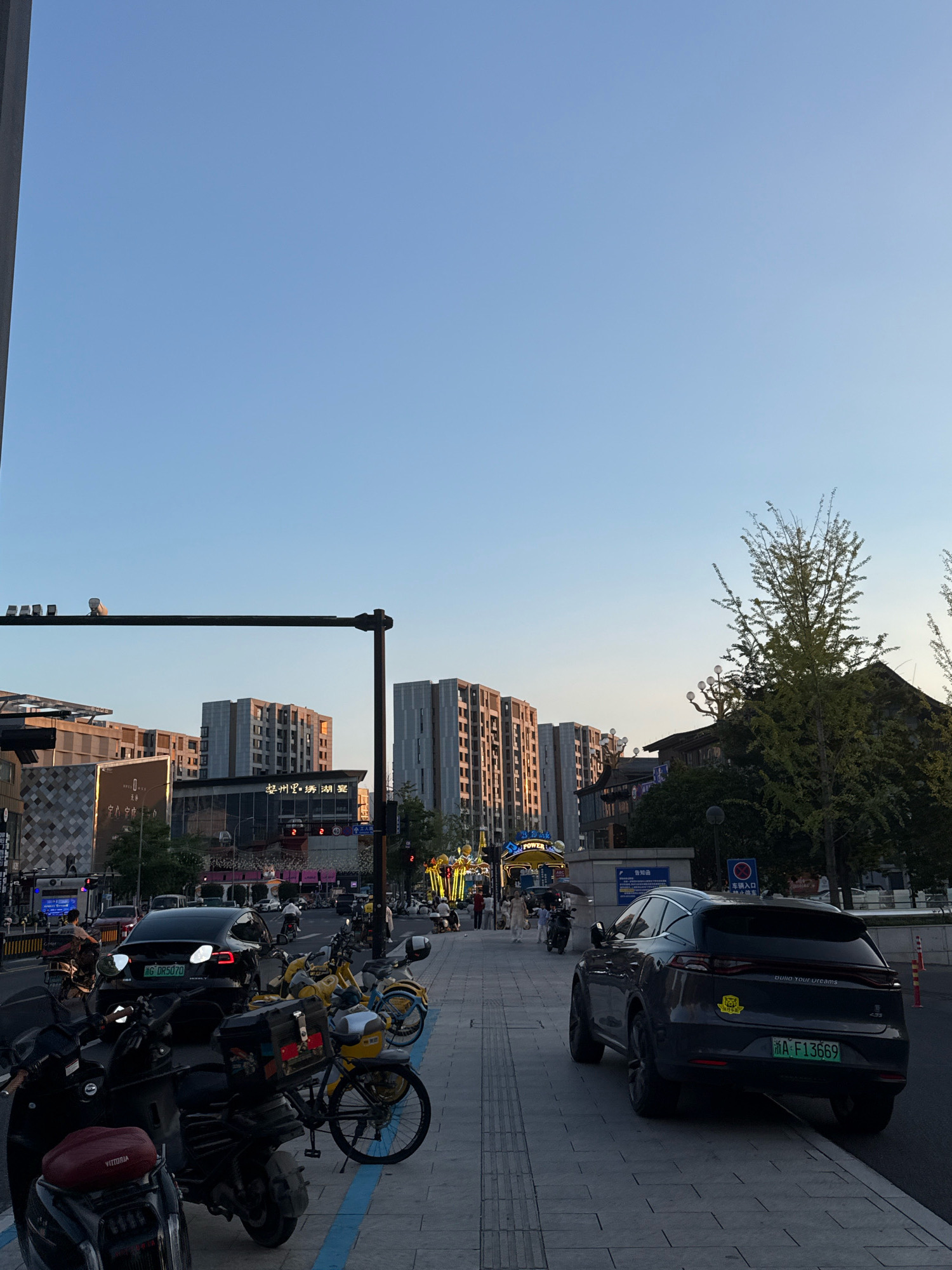 The image depicts a street scene in Yiwu, China, during what appears to be late afternoon or early evening, given the warm, low sunlight illuminating the buildings. The sky is clear and blue.
In the foreground, a sidewalk is lined with parked motorcycles and bicycles, predominantly dark in color with some yellow accents. A dark-colored SUV is parked on the right side of the road, facing away from the viewer. Its license plate is visible and reads "A·F13669". A sticker is affixed to the rear windshield.
The street itself has a few vehicles, including a black Tesla with its taillights illuminated. Traffic lights are visible on poles. Pedestrians are present on the sidewalk, some walking and others appearing to be interacting with the parked vehicles. One pedestrian is holding an umbrella.
The background features modern multi-story buildings, some with large windows reflecting the sunset. One building has prominent Chinese characters. Trees, including a large deciduous tree with spreading branches, are scattered throughout the scene. Streetlights and lampposts are also visible. The overall impression is that of a busy urban environment.