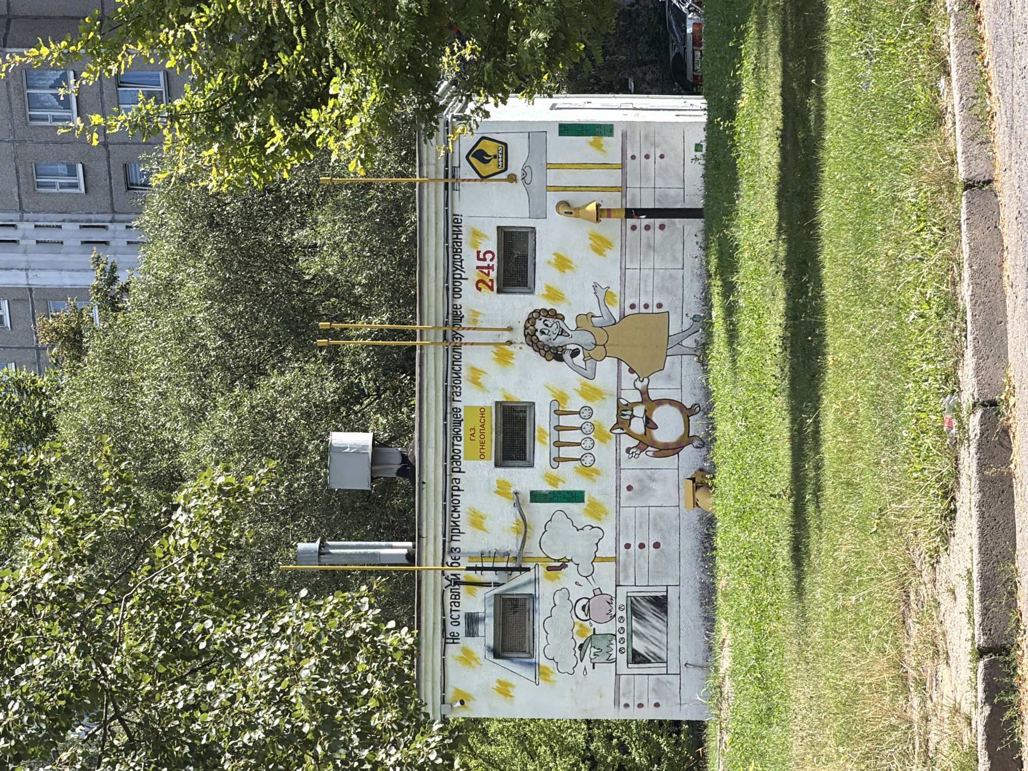 A small, white, rectangular building with a mural on its facade is situated in an urban environment, likely in Minsk, Belarus, as indicated by the presence of Cyrillic text. The building appears to be a gas distribution point, evidenced by the prominent yellow gas pipes and a gas safety symbol.
The mural depicts a whimsical scene, featuring a cartoonish woman with curly hair and a yellow dress, a cartoon cat, and kitchen elements like a stove, pots, and utensils. The woman is depicted with a surprised or animated expression. The text on the building, written in Russian, reads "Не оставляй без присмотра работающее газоиспользующее оборудование!" which translates to "Do not leave gas-using equipment unattended!" and "ГАЗ ОГНЕОПАСНО" meaning "GAS IS DANGEROUS". A number "245" is also visible on the building, possibly an identifier.
The building is surrounded by green grass and trees, with a larger, grey apartment building visible in the background, characteristic of Soviet-era architecture. The lighting suggests it is daytime, with shadows cast on the grass, indicating sunlight. The overall scene presents a contrast between the industrial function of the building and the playful, artistic mural intended to convey a safety message.