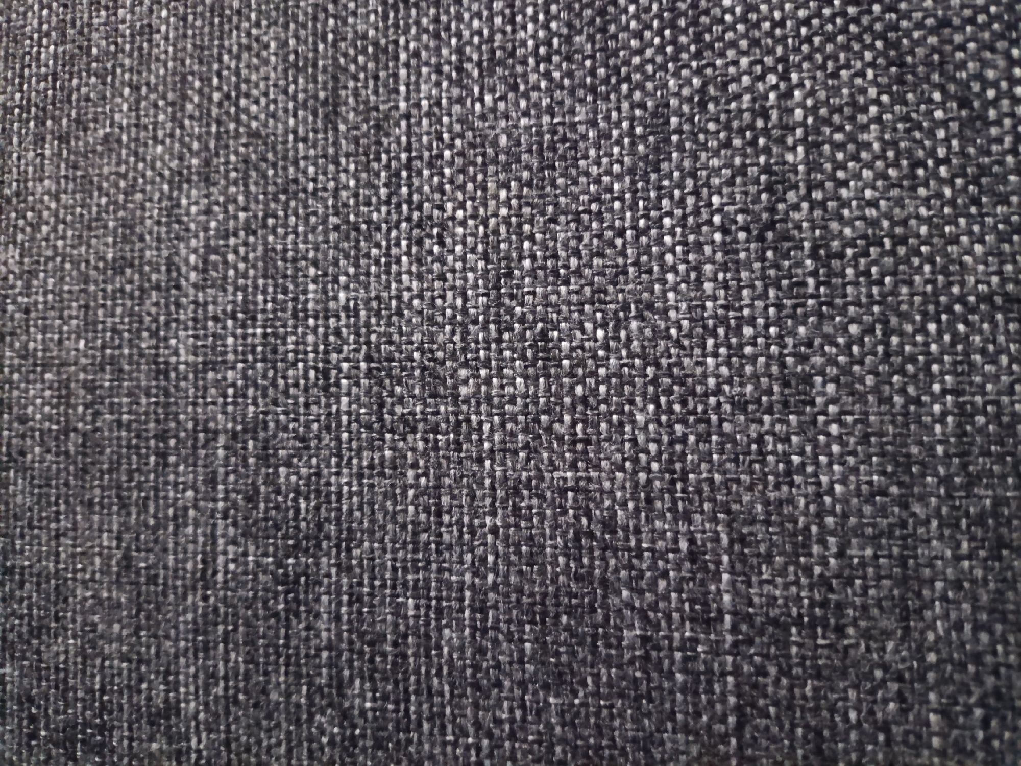 The image is a close-up, overhead shot of dark gray woven fabric. The texture of the fabric is clearly visible, with individual threads interlocking to create a textured surface. The lighting is even, with no strong shadows, highlighting the intricate weave pattern. The color is a uniform dark gray, with subtle variations in shade due to the texture of the threads. There are no people, objects, or environmental features present in the image. It is a still life shot focused entirely on the textile.