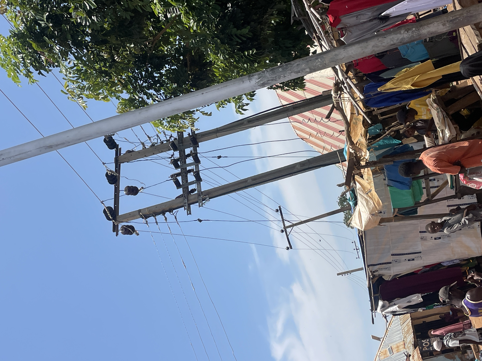 This image depicts an outdoor market scene, likely in Zalau, Nigeria. Several makeshift stalls, constructed from wood and sheeting, are visible, displaying a variety of clothing items hanging on racks. People, including men and boys, are present, some interacting near the stalls, while others appear to be vendors or customers. In the background, power lines stretch across a bright blue sky with scattered clouds. A large green tree with dense foliage is also visible, partially obscuring some of the stalls and power lines. The overall impression is one of a bustling, informal marketplace with basic infrastructure.