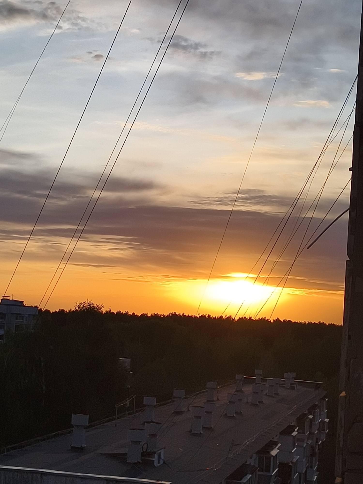The image shows a sunset viewed from a high vantage point in an urban setting. The sky is a mix of light blue, gray, and orange hues, indicating the sun is setting or has recently set. Power lines crisscross the upper portion of the frame. Below the horizon, a line of trees is visible, followed by a darker area. In the foreground, the top of a building with several ventilation units is visible, and part of a building facade is present on the right side of the image.