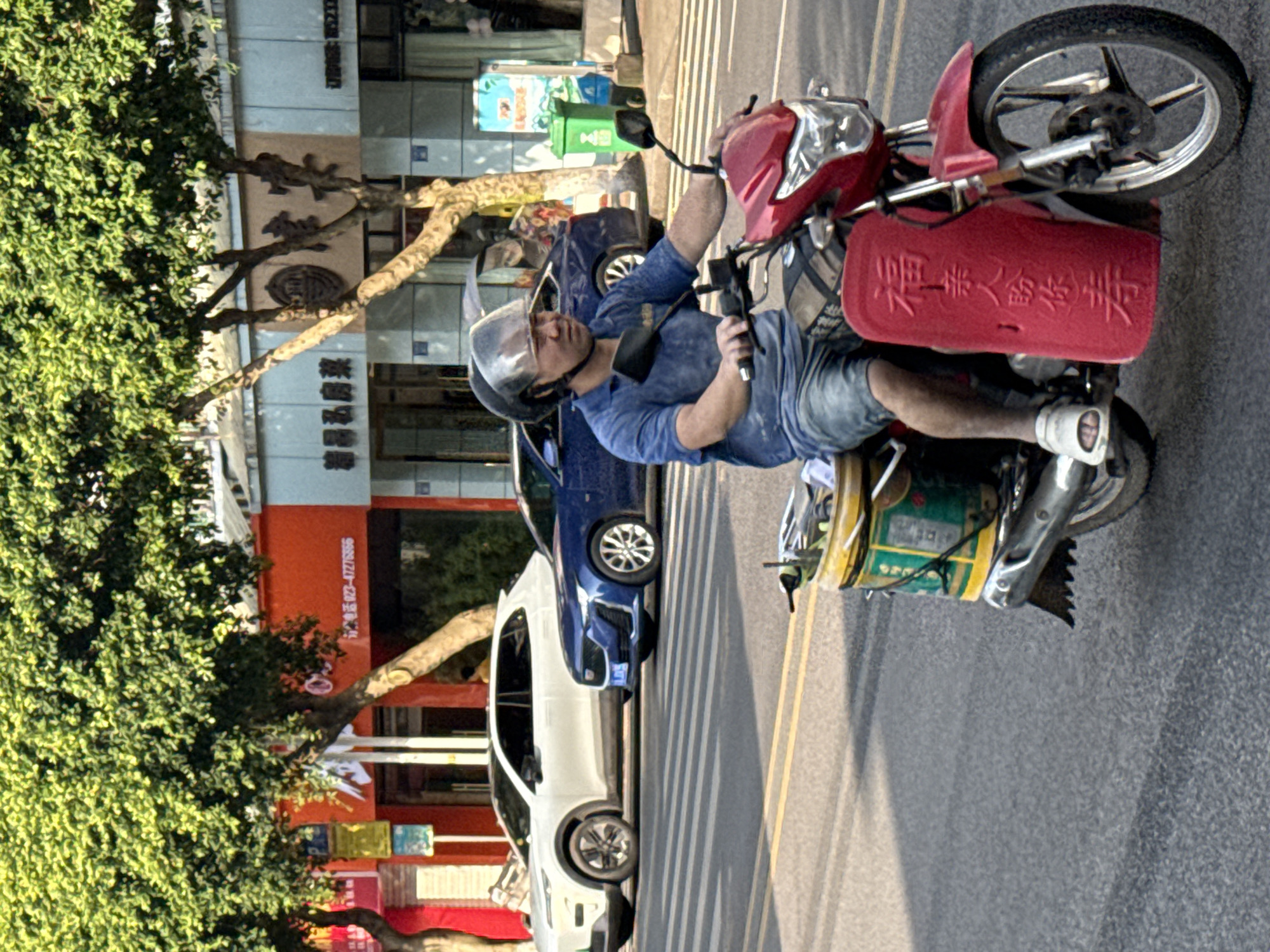 An upright-oriented shot captures a man on a red scooter traveling or paused on a sunlit street in Jiangjin, China. The man, wearing a blue short-sleeved shirt, denim shorts, and white sneakers, has a helmet with a clear visor on his head and appears to be focused forward. His right hand rests on the scooter's handlebar.
The red scooter has a prominent red plastic box attached to its front, featuring embossed Chinese characters that read "福泽人安泰事" (Fú zé rén ān tài shì), roughly translating to "Blessings bring peace and prosperity to people." A green and yellow container or basket is visible on the side of the scooter.
In the background, a row of buildings lines the street, partially obscured by lush green trees. A white car and a blue car are parked or moving on the left side of the road, behind the trees. The street itself has clear double yellow lines marking its center. Shadows are cast across the asphalt, indicating bright daytime sunlight. On one of the red buildings, a phone number "023-67278556" is partially visible. Another building appears to have Chinese characters, with "茶" (chá, meaning tea) and "馨阁" (xīn gé, meaning cozy pavilion) partially discernible. Blue and green trash bins are also visible in the background, near the buildings.
