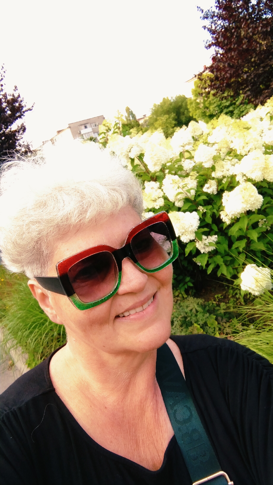 A mid-shot selfie shows a woman with short, white hair and a tan complexion. She is wearing large, square sunglasses with black frames accented with red and green. She is smiling and looking slightly to her right. She has on a black t-shirt, and a dark green strap with a raised pattern is visible across her chest. The background is a garden setting with lush green foliage, white flowers, and a dark red tree on the right. In the distance, a light-colored building with several windows is partially visible. The overall impression is of a bright, sunny day. The photograph was taken in Horishni Plavni, Ukraine.
