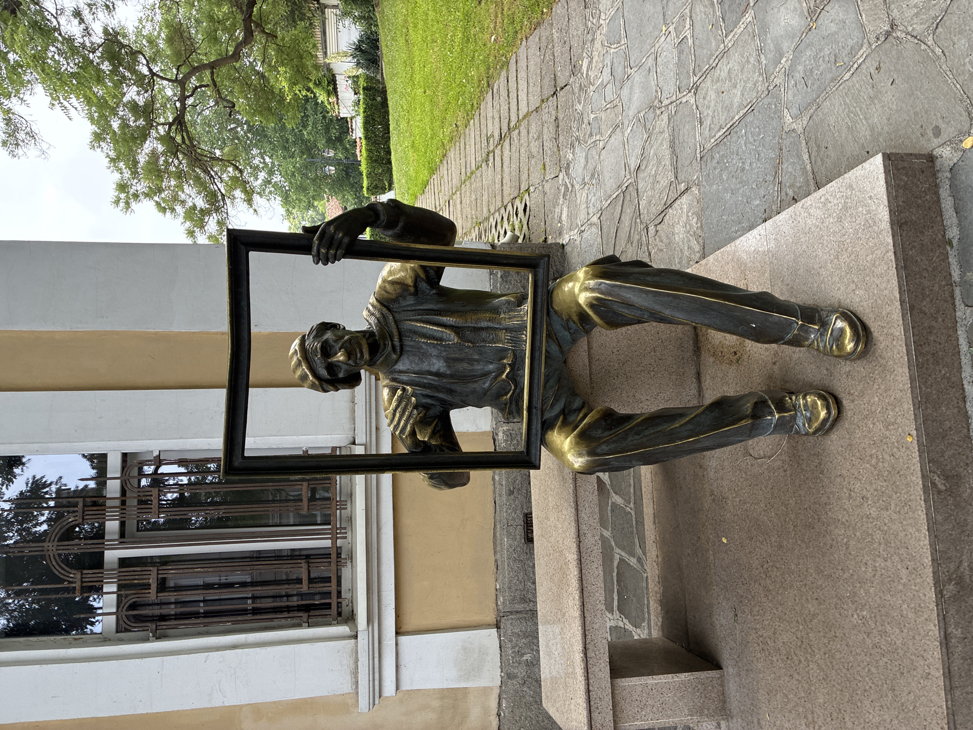 The media file displays a bronze statue of a man, positioned outdoors in Plovdiv, Bulgaria. The statue, depicting a figure that appears to be an artist wearing a cap or beret, is shown seated or kneeling on a stone plinth. The man holds an empty, rectangular, dark-colored frame vertically in front of him, looking through it or presenting it. Some parts of the statue, such as the shoes, trousers, and elements of the frame, have a polished, golden finish, contrasting with the darker bronze of the rest of the figure.
The setting includes a paved walkway made of irregular stone slabs to the right of the statue, bordered by green grass and trees in the background. To the left, behind the statue, is the facade of a building with light-colored walls and windows featuring decorative, dark metal grilles. The lighting suggests it is daytime with a clear sky, visible through the foliage of the trees. There is no visible text in the image. The overall scene conveys a sense of contemplation or artistic presentation within a public outdoor space.