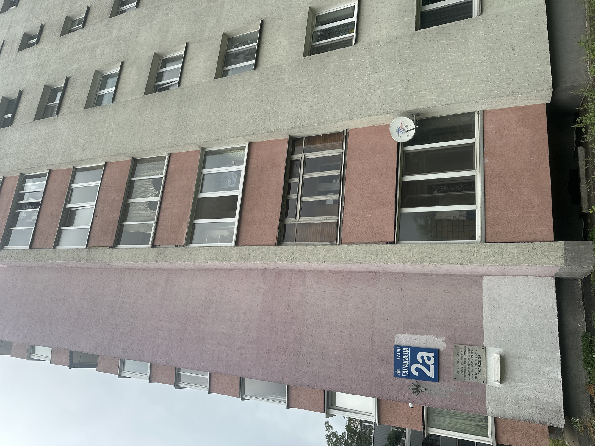 The image shows a residential building located at Galadzyeda Street, 2a, in Minsk, Belarus. The building has a distinctive architectural design with a pink and gray facade. Several windows and balconies are visible on the building's exterior. A blue street sign indicating "ВУЛІЦА ГАЛАДЗЕДА 2а" (Galadzyeda Street 2a) is mounted on the pink part of the facade. Below the street sign, there is a memorial plaque with text in Belarusian. A satellite dish is attached to the facade near one of the windows. The sky appears overcast.