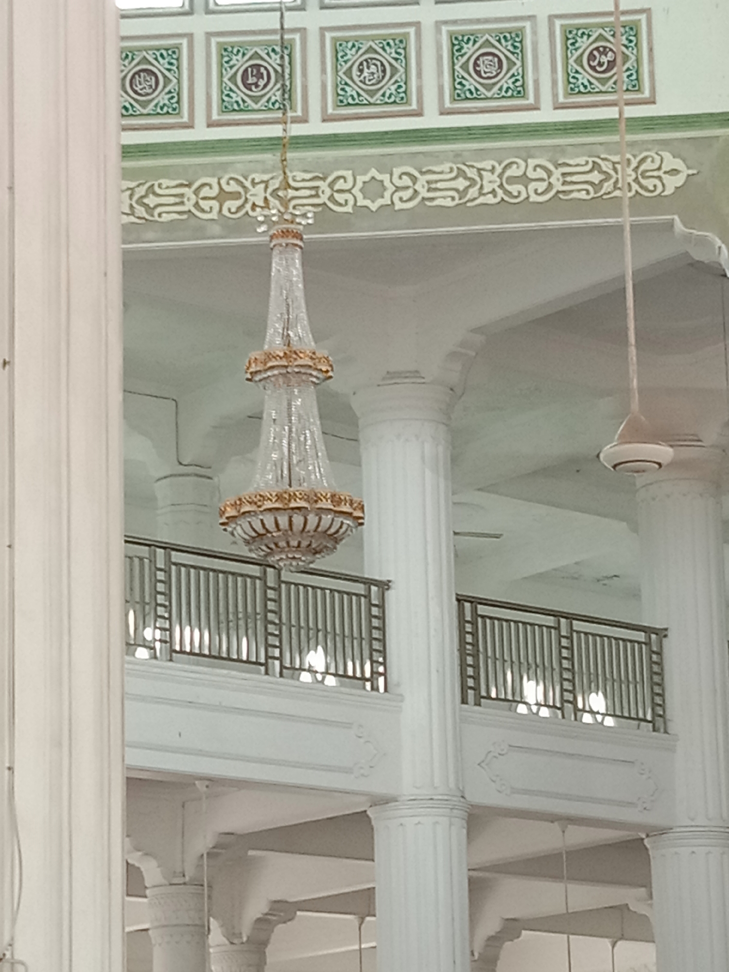 This is an interior shot of the Baiturrahman Grand Mosque in Lhokseumawe, Indonesia, focusing on its ornate ceiling and decorative elements. A large, multi-tiered crystal chandelier with gold accents hangs prominently in the center. Above the chandelier, the ceiling features square panels adorned with intricate green and white Islamic calligraphy and geometric patterns. A decorative frieze, also in white and green, runs along the upper wall. Large white columns support the vaulted ceiling, and a metal railing, part of an upper level or gallery, is visible in the mid-ground. A white ceiling fan is also present. The lighting is soft, suggesting diffused natural light or interior illumination. There are no people or animals visible in the frame.