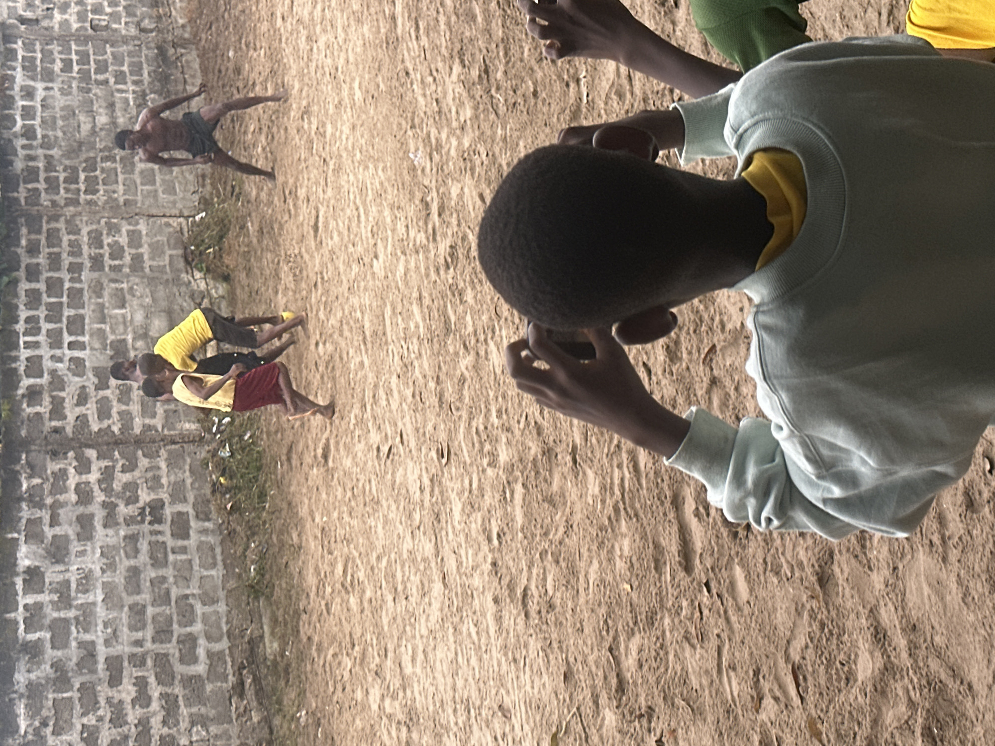 In a sandy outdoor area with a rough brick wall in the background, several young Black individuals are engaged in a game of football. In the foreground, a young Black person, facing away from the camera, is wearing a light blue sweatshirt over a yellow shirt and is holding a smartphone, appearing to record the event. Behind this person, a group of three other young Black individuals are actively playing. One is wearing a yellow tank top and red shorts, another is in a yellow t-shirt and dark shorts, and a third, shirtless, is wearing dark shorts. The ground is composed of sand and dirt, with some sparse vegetation visible near the wall. The environment suggests a casual, informal setting, possibly a neighborhood playground or open space within the city of Badagry, Nigeria.