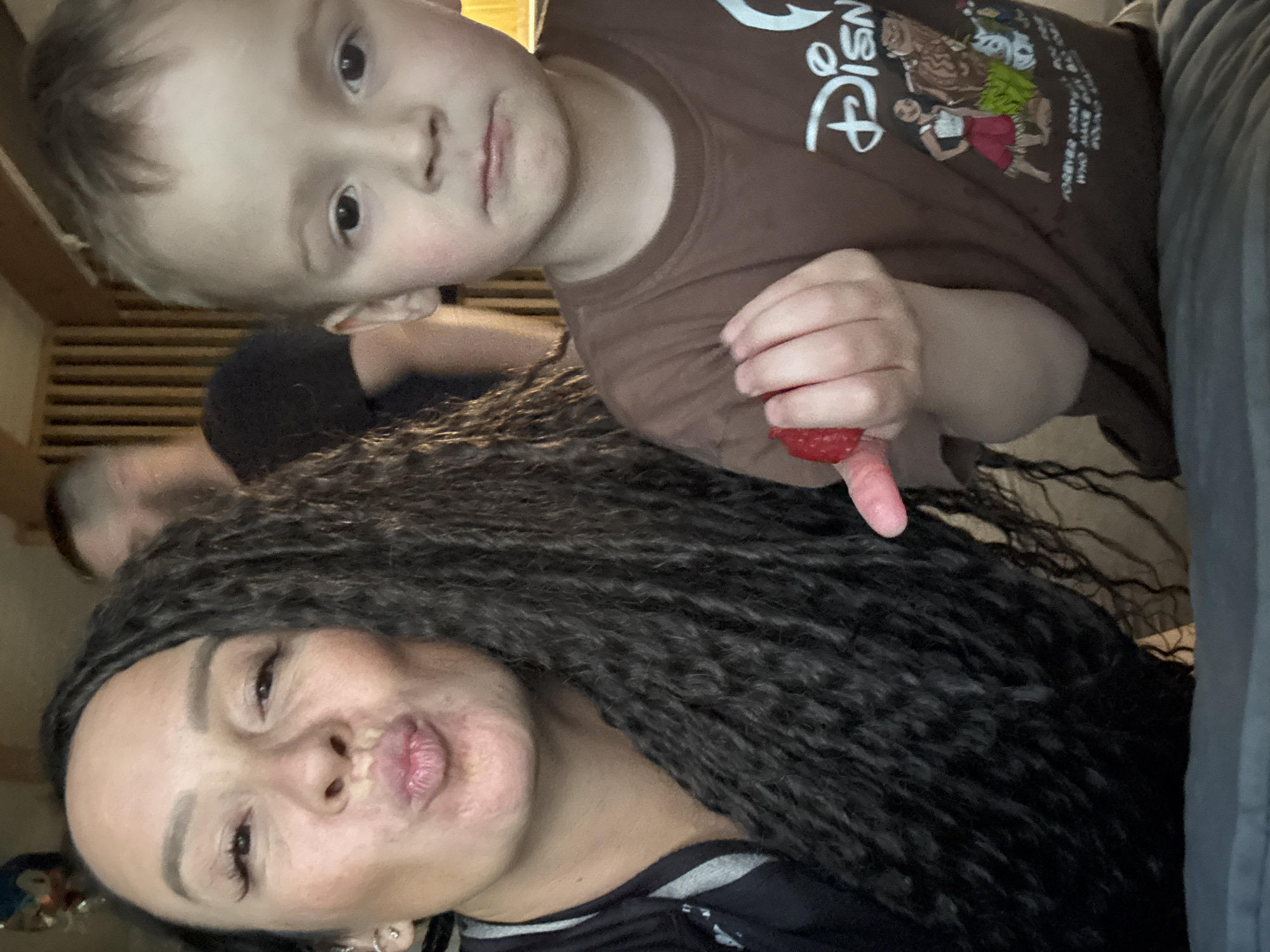 The original image is a vertically oriented selfie, captured indoors. It primarily features a woman and a young child, with a third person partially visible in the background.
In the foreground, a woman with long, dark, braided hair looks towards the camera, making a playful pouting or kissing face with her lips. She has visible makeup, including defined eyebrows and eyelashes, and wears a dark top with a grey accent and a small earring.
To the upper right of the woman is a young child with short, light-colored hair, looking directly at the camera with a neutral expression. The child is wearing a brown t-shirt featuring a white "Disney" logo and cartoon characters, specifically characters from the movie "Moana," including Maui and Moana herself. The text "FOREVER CRAZY WHO ARE YOU" is partially visible below the characters. The child is holding a red strawberry in their right hand, with one finger extended beneath it.
In the blurred background, to the left of the child and behind the woman, the head and shoulder of another person with short, dark hair are visible, appearing relaxed. The setting is an indoor space, suggested by a wooden slatted structure visible in the background, possibly part of a wall or furniture. The scene conveys a casual and familial moment. No specific time of day or weather can be determined from the image. The provided location context is Chaikovsky, Russia.