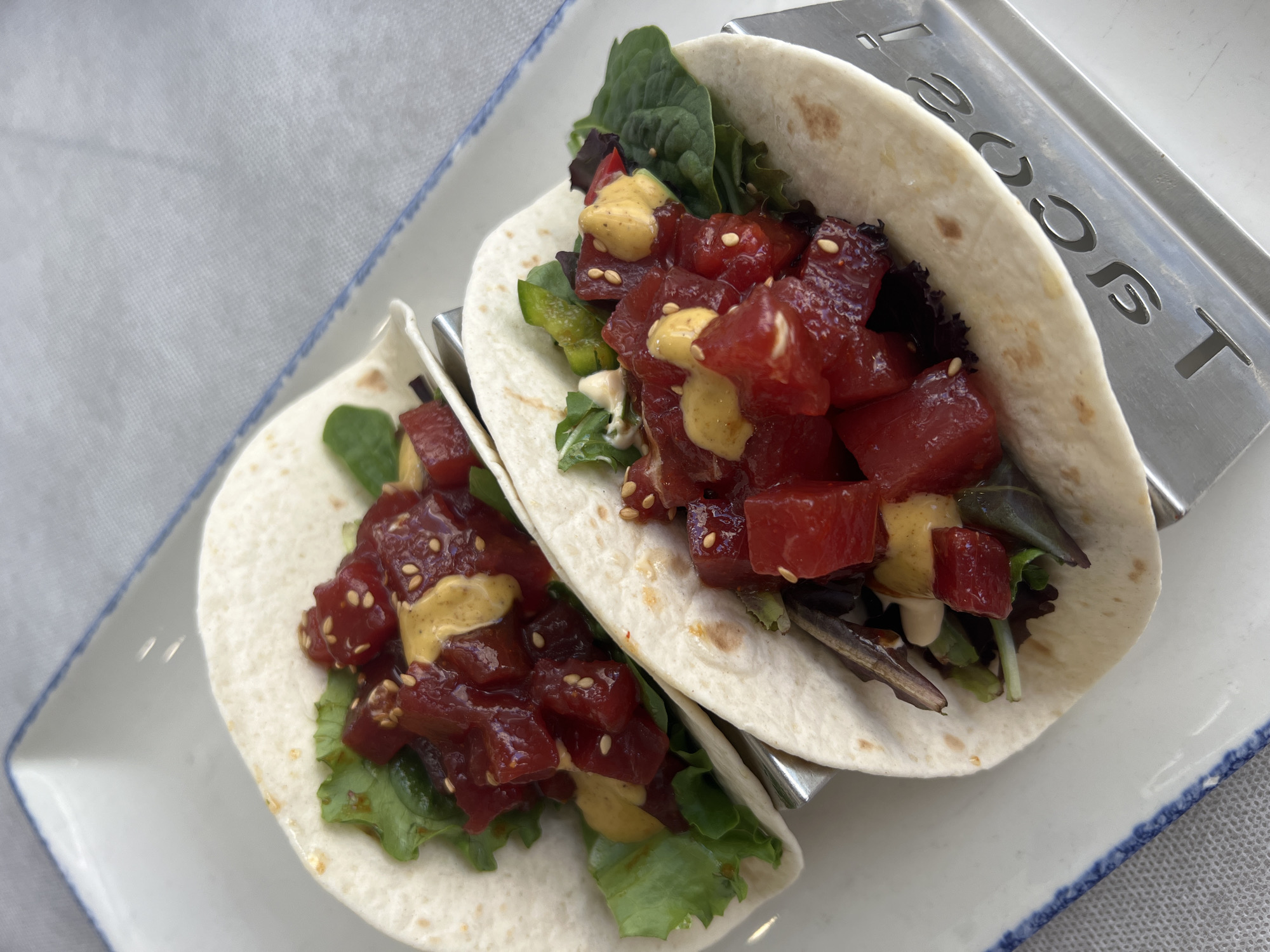 Two tuna poke tacos are presented on a white rectangular plate with blue trim, resting on a textured grey tablecloth. Each taco is made with a soft, white tortilla. The tacos are filled with diced raw tuna, green lettuce, and a drizzle of a yellowish sauce, possibly a spicy mayo or aioli. Sesame seeds are sprinkled over the tuna. The tacos are held upright in silver metal taco holders, one of which is partially visible and has the word "Tacos" cut out of it. The background is softly blurred, suggesting a close-up shot focused on the food. The scene appears to be indoors, likely a restaurant setting, in Valdeconejos, Spain.
