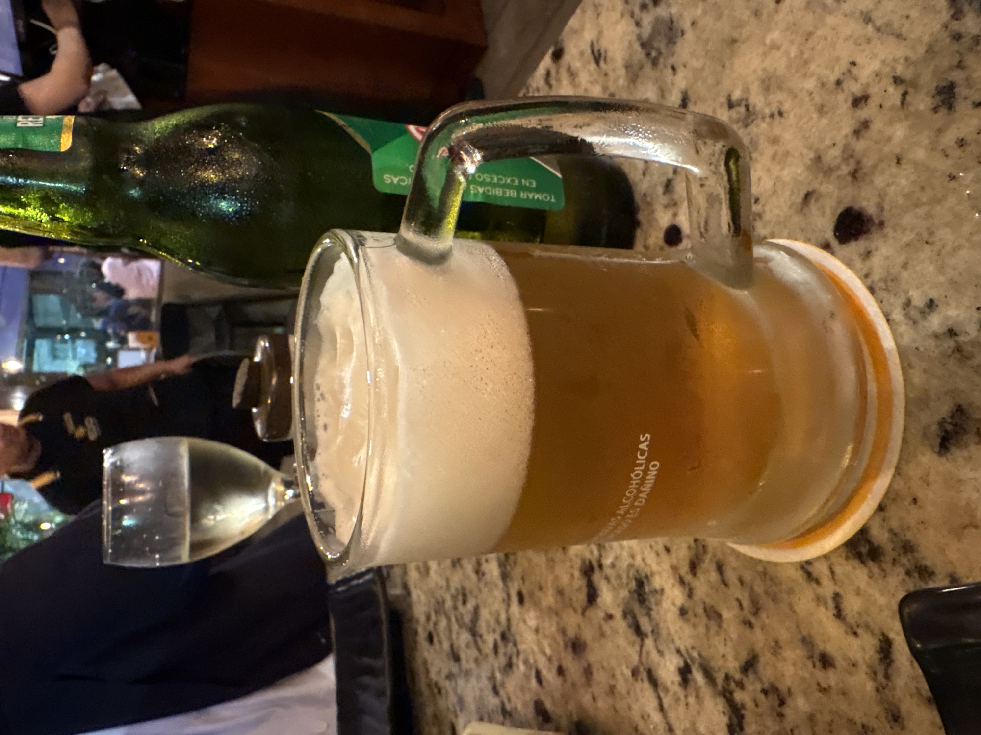 The image shows a close-up of a beer mug filled with amber-colored beer and topped with a thick layer of foam. The mug has a handle and is placed on a granite-like countertop. To the right of the mug is a condensation-covered green glass bottle of beer, with a partially visible label. In the background, out of focus, are several people and an outdoor setting, possibly a restaurant or bar, with tables and chairs. The ambient lighting suggests it is daytime or early evening. The text on the beer mug, in white, reads "IS ALCOHÓLICAS" and "ES DAÑINO," which translates to "ARE ALCOHOLIC" and "IS HARMFUL." The label on the beer bottle contains the words "TOMAR BEBIDAS" and "EN EXCESO," translating to "DRINK BEVERAGES" and "IN EXCESS." Based on the presence of these specific warning messages and the typical cultural context of such establishments, this scene is highly likely to be in Magdalena del Mar, Peru, a district known for its social venues and restaurants.