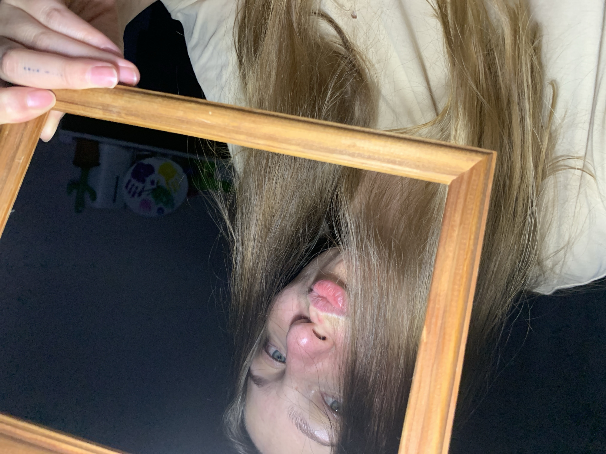 A young individual with long, light brown hair is captured in a close-up, slightly tilted portrait. The person is holding a light-brown wooden picture frame, presenting their face through its opening. Their light-colored eyes gaze directly at the viewer, and their lips are pursed into a distinct kissing or pouting gesture, appearing pink and slightly glossy. The nose shows a reddish tint. Hair frames the face, partially obscuring the left side. The individual wears a light-colored, possibly cream or off-white, garment. In the lower right, a hand holds the frame, revealing manicured fingernails painted pink, and a small tattoo consisting of four blue dots on the ring finger. The background is dimly lit and dark grey or black, with a slightly textured appearance. To the right, a white decorative plate with colorful handprints and a small green potted cactus are visible on a surface, suggesting an indoor setting. The image contains no discernible details indicating the location as Chaikovsky, Russia.