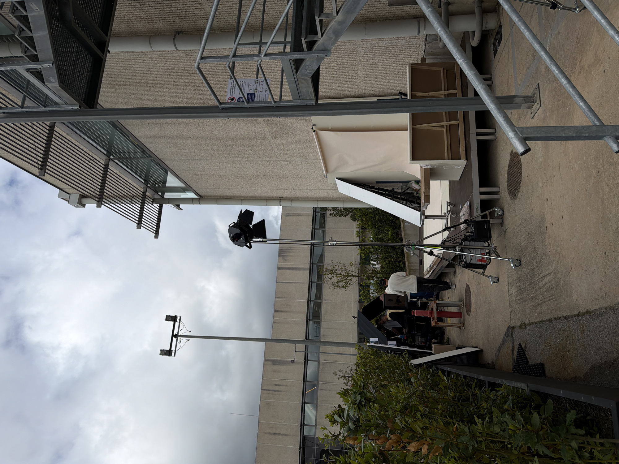 The image shows an outdoor film set. A large industrial building with multiple rows of windows is visible in the background, with film equipment set up in front. This includes lights, reflectors, and cameras on tripods. Several people are present, appearing to be crew members working on the set. A cloudy sky can be seen. The location in the city of Fibracolor, Spain, is not evident from visual cues.