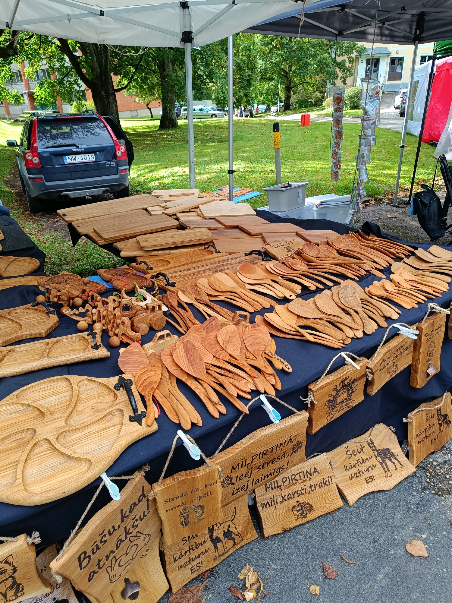 This is a medium-shot, eye-level outdoor photograph of a market stall selling wooden crafts, likely in Kazdanga, Latvia. The stall is set up under a white tent, with trees and a building in the background, suggesting a public park or gathering space.
Dominating the foreground and midground are numerous wooden items, primarily kitchenware and decorative pieces. There are stacks of wooden cutting boards, a large collection of wooden spoons and spatulas, and several intricately carved wooden toys, including a caterpillar-like vehicle and what appear to be small animal figures. Some of the items have metal handles.
Hanging from the stall are small wooden signs with etched text and images, including animal silhouettes and decorative motifs. These signs are attached with rope and some are secured with clothespins.
In the background, partially visible under the tent, is a dark blue Volvo SUV with a license plate reading "NW-4019". Beyond the stall, a grassy area with trees is visible, and in the further distance, buildings and other parked vehicles can be seen. The ground is a mix of asphalt and fallen leaves, indicating an autumn setting. No people are prominently featured in the image.