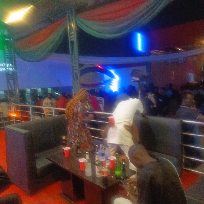 The scene depicts a gathering, possibly at a club or lounge. A woman with long, braided hair wears a floral dress and stands near a table cluttered with red plastic cups and various bottles. A man in a white outfit hugs another man from behind, who also holds a red cup. Another man is sitting in the foreground. Behind them, a railing separates the area from a dance floor with blue and white lighting. Draped fabric in green, white, and orange adds a decorative touch.