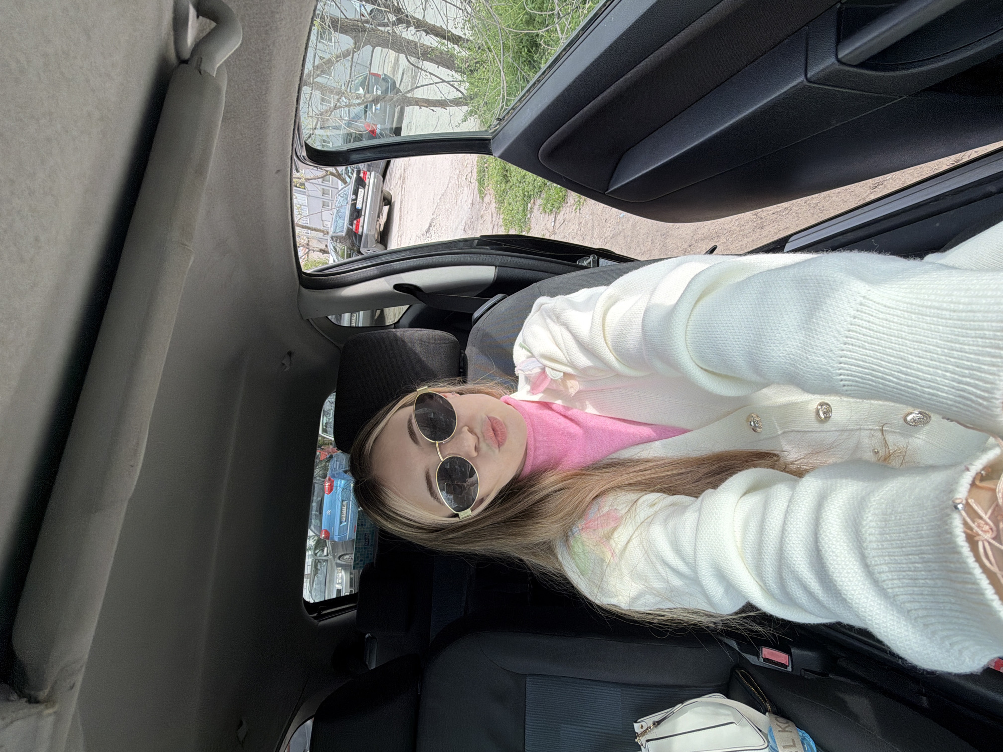The image shows a young woman inside a car, presumably in Plovdiv, Bulgaria, judging by the visible license plates of the cars outside. She is wearing sunglasses, a pink top, and a white cardigan. The car door is open, revealing a street scene with parked cars and trees. The interior of the car is visible, including the dashboard, seats, and a white purse.