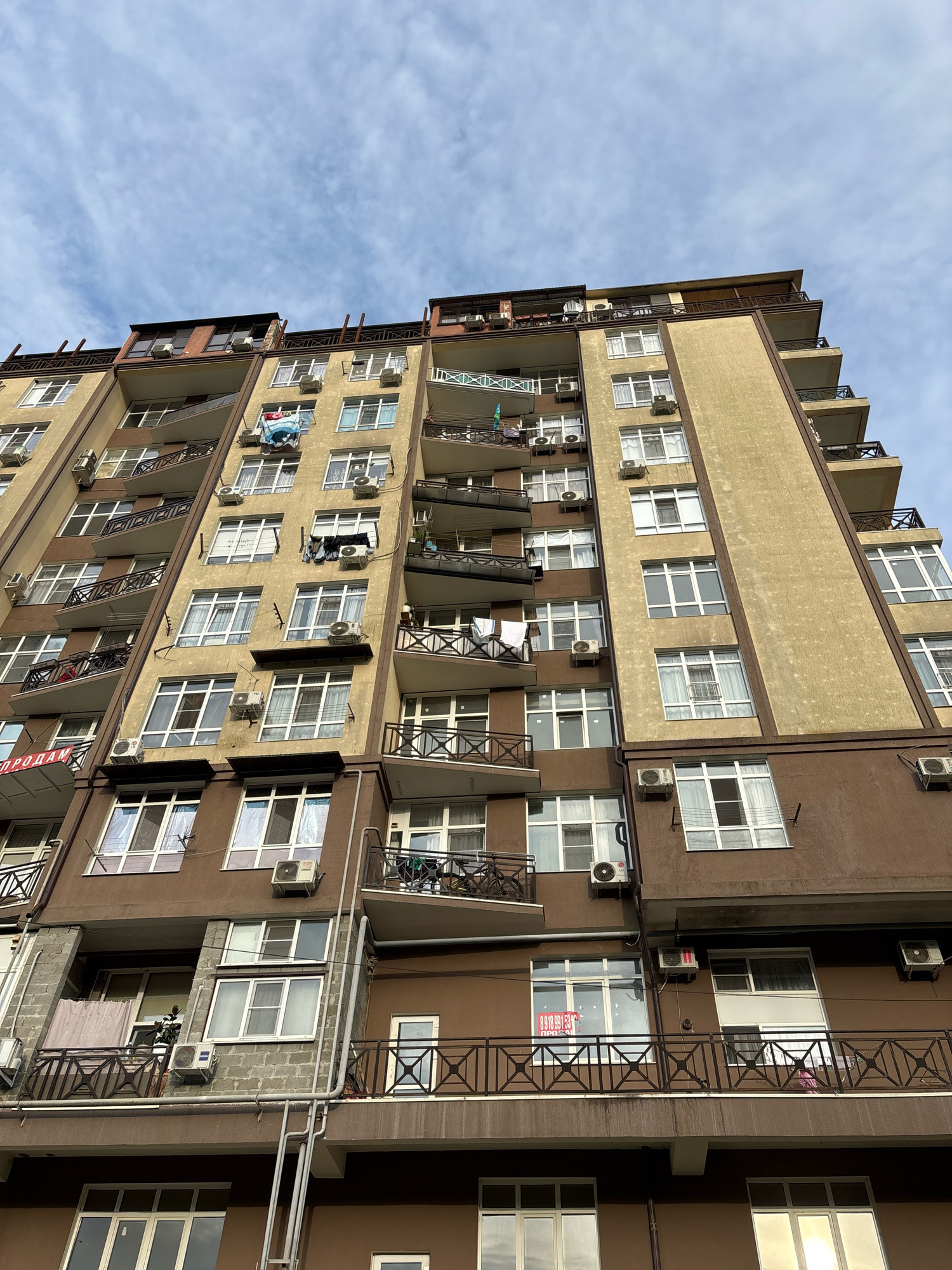 The photograph shows a tall apartment building in Sochi, Russia, with a facade of brown and beige tones. The building features numerous balconies with dark railings and many windows. Several air conditioning units are visible mounted on the exterior walls. Laundry is hanging on some of the balconies. The sky above is cloudy with patches of blue. The perspective is from a low angle, looking up at the building.