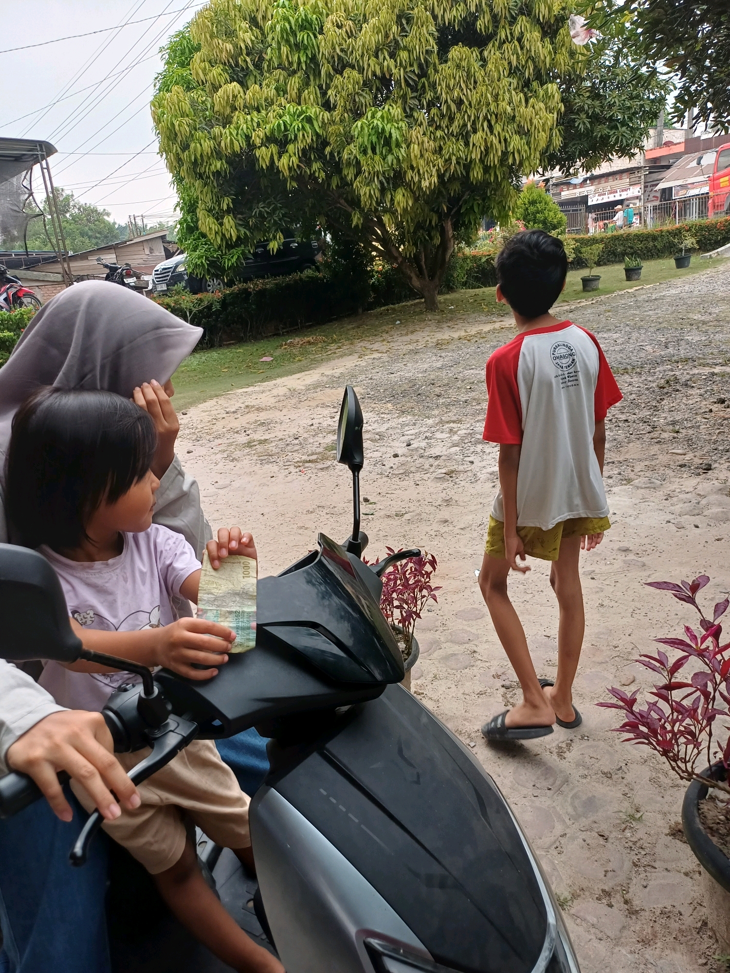 The image shows a street scene in Dumai City, Indonesia. In the foreground, a child wearing a light purple t-shirt sits on a motorcycle and holds a 1000 Indonesian Rupiah banknote. Next to the child, a person wearing a grey hijab is also on the motorcycle. The child is looking to the left. Behind them, a young boy with dark hair stands in a red and white t-shirt and yellow shorts. He is looking away from the camera towards a large tree and a row of shops in the background. The ground is covered in gravel and dirt. There are several potted plants with red leaves visible. In the background, there are buildings, parked motorcycles, and cars, suggesting a residential or commercial area. The sky is overcast.