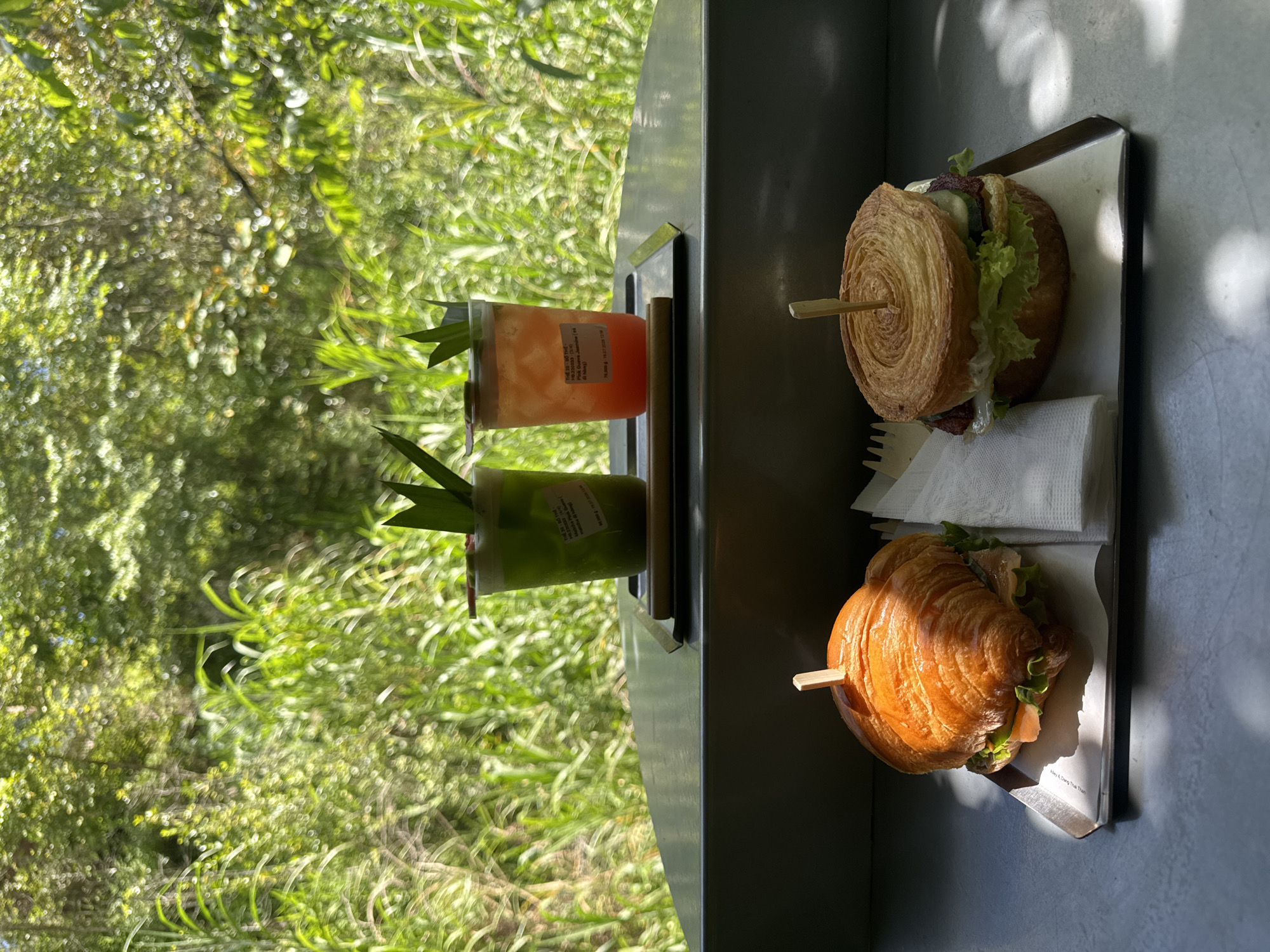 The image shows a close-up of a tray with two food items and two drinks placed on a surface in front of a blurred background of lush greenery, characteristic of Da Lat, Vietnam.
In the foreground, on the left, is a croissant sandwich filled with what appears to be salmon and lettuce. A wooden skewer is inserted into the top of the croissant. To the right of the croissant sandwich is another sandwich, this one with a circular, layered bun, also secured with a wooden skewer. This sandwich appears to contain lettuce, cucumber, and other fillings. Both sandwiches are on separate paper trays. A white napkin is placed between them.
In the background, on a slightly elevated surface, are two clear plastic cups containing colorful drinks. The cup on the left holds a green beverage, possibly a matcha or herbal drink, garnished with a leaf. The cup on the right contains an orange-red beverage, also garnished with a leaf. Both cups have labels adhered to them. Behind the drinks, the blurred background of dense, green foliage suggests an outdoor or semi-outdoor setting with abundant natural vegetation, typical of Da Lat's landscape. The lighting suggests a bright, sunny day, with sunlight casting distinct shadows on the surface where the food is placed.