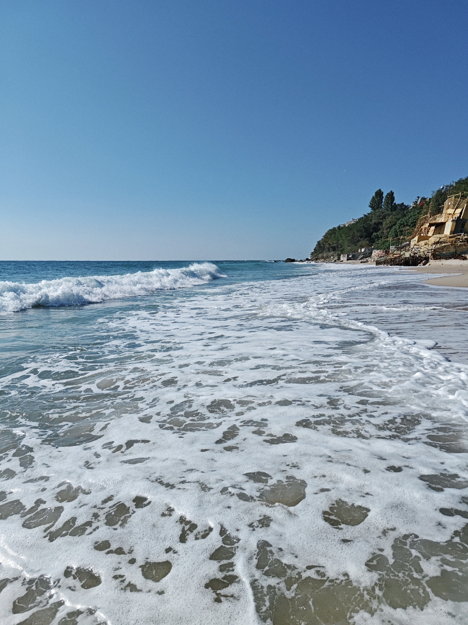 A sun-drenched beach on a clear day, with the turquoise sea meeting the sandy shore. Gentle waves create frothy white foam that stretches towards the foreground, revealing the sandy seabed beneath. In the background, a verdant hillside rises, topped with dense green trees. Along the hillside, weathered, ochre-colored structures are visible, hinting at past construction or ruins. The sky is a vast expanse of uninterrupted blue. No people are present in the scene.
