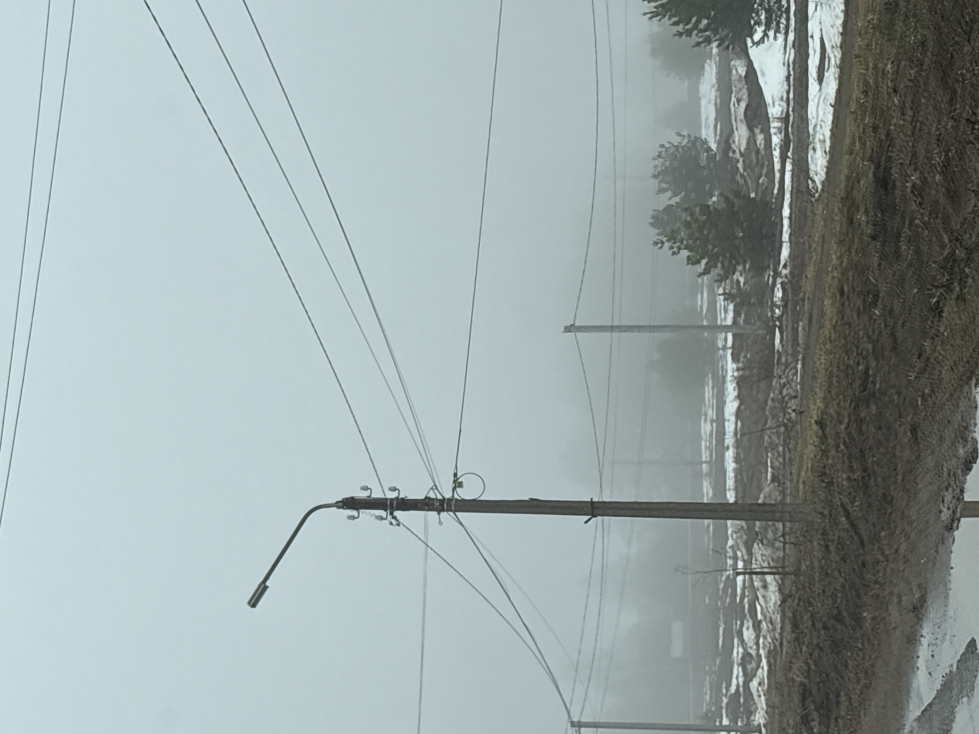 The image depicts an overcast, possibly foggy or snowy, outdoor scene in Прикамский, Russia, evidenced by the presence of snow and the typical infrastructure. A utility pole with a streetlight is prominently featured in the foreground. Power lines crisscross the sky. A blurred landscape is visible in the background, showing signs of a rural environment with trees and fields. The ground is covered in a mix of snow and vegetation. No people are visible.