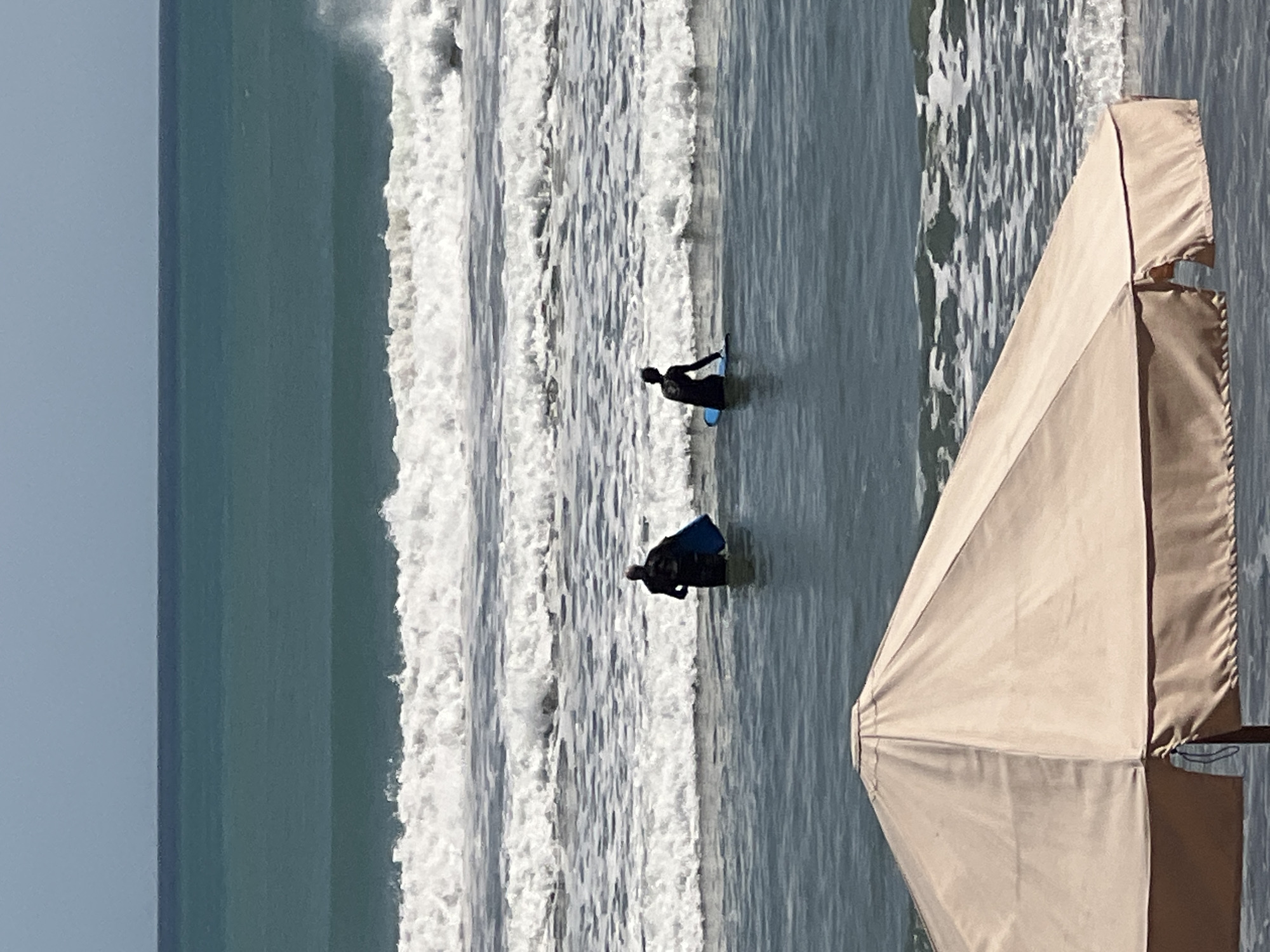 Two individuals, each holding a blue surfboard, stand in the ocean near Kuta, Indonesia. The person on the left is wearing a dark wetsuit and the person on the right is also in dark attire. They are surrounded by several rows of waves, with white foam breaking on their crests. The water is a deep teal color, transitioning to a lighter blue towards the horizon. In the foreground, a portion of a large, tan beach umbrella is visible, its fabric taut and casting a shadow. The sky above is a pale, clear blue.