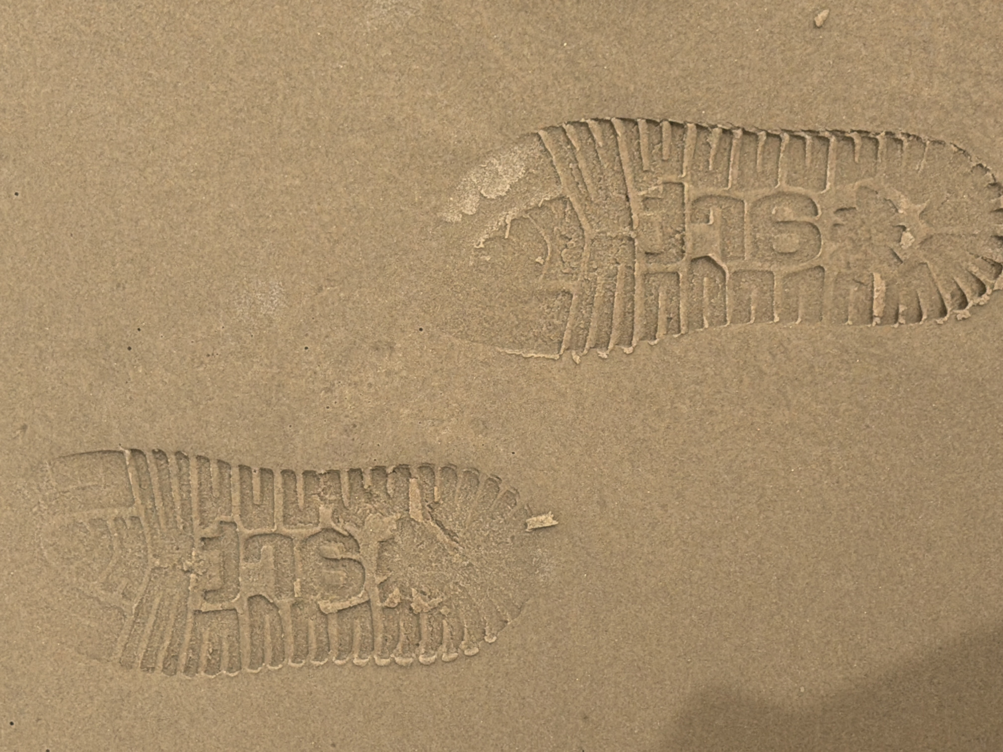 Two shoe prints are visible in a sandy or dusty substrate. The shoe tread appears to have a distinctive pattern with prominent lettering or characters embedded within it. The surrounding environment appears to be a flat, textured surface. The location is not discernible from the image.
