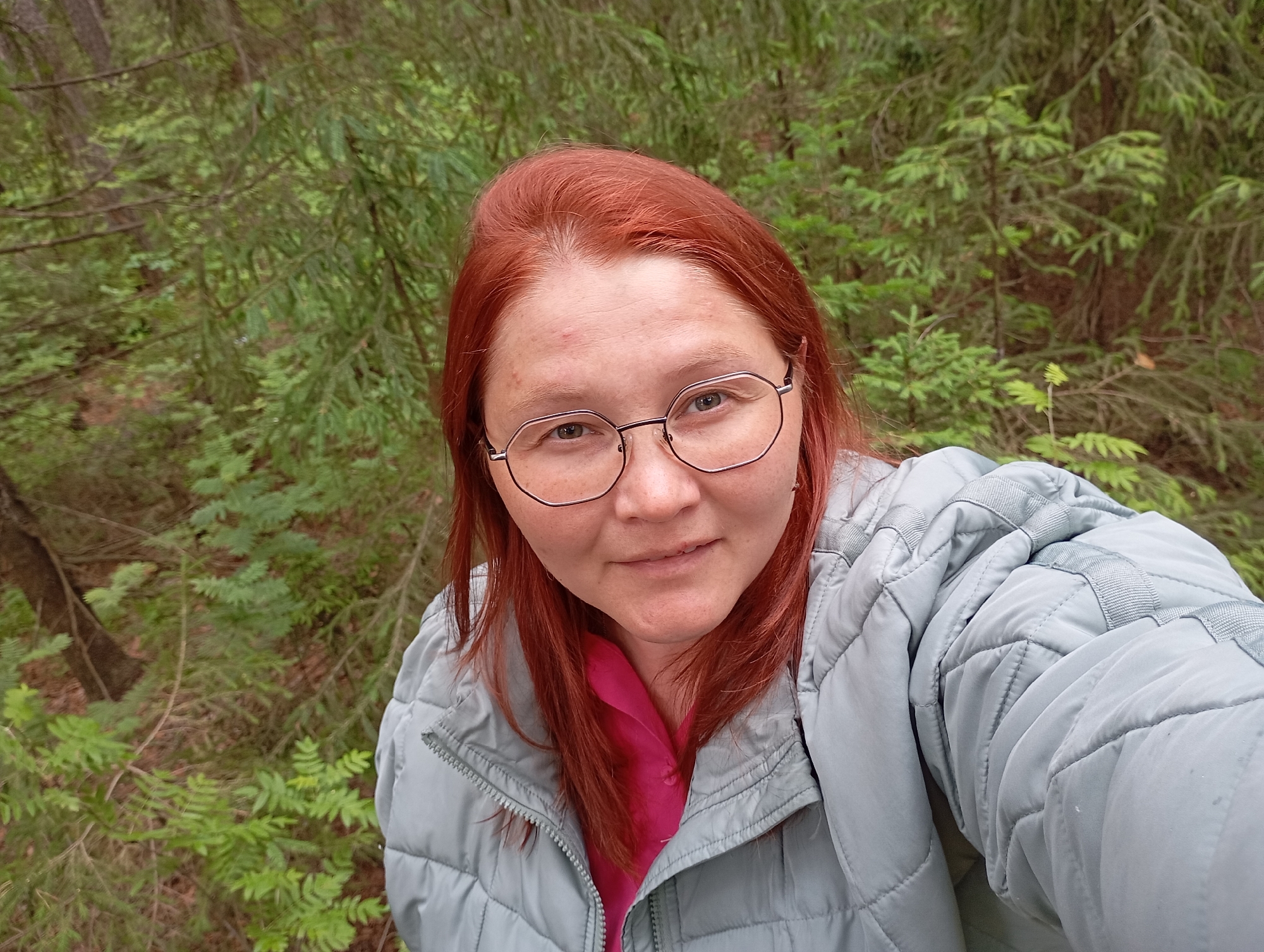 A close-up outdoor selfie captures a woman with distinct red hair and light-colored skin, wearing modern hexagonal-framed glasses. She looks directly at the camera with a neutral expression, possibly a slight, subtle smile. Minor skin imperfections are visible on her forehead and nose.
She is dressed in a light grey or light blue quilted jacket, which is partially unzipped to reveal a bright pink collared shirt underneath. The jacket features a zippered front and padded sections, suggesting cool or mild weather.
The background is a dense, verdant forest, filled with various shades of green foliage. Coniferous trees, possibly spruce or fir, dominate the backdrop, alongside lighter green undergrowth. The background is softly blurred, emphasizing the subject in the foreground.
The lighting appears to be natural daylight, likely diffused by an overcast sky or the forest canopy, as there are no harsh shadows. The image depicts a casual moment in a natural, wooded environment. No visible text is present in the image. The setting is consistent with a forest in Chaikovsky, Russia.