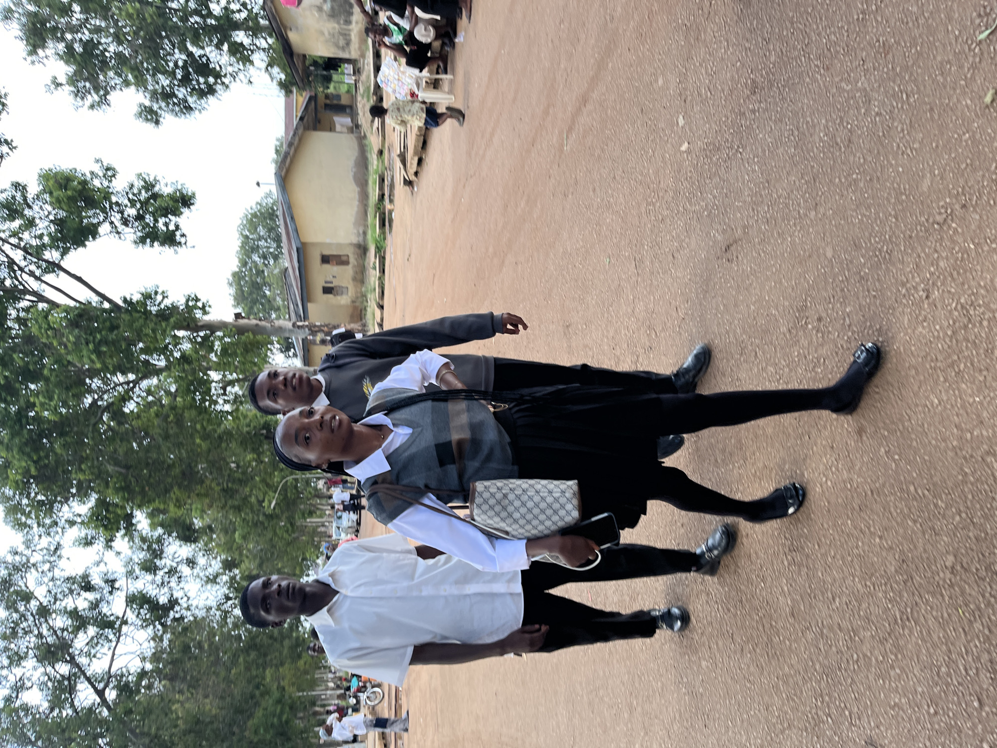 Three young individuals, likely students in uniform, are captured outdoors in what appears to be a street or pathway in Rafin Par, Nigeria. The image is oriented vertically but depicts the subjects standing upright.
In the foreground, a young man on the left wears a white short-sleeved button-up shirt and dark trousers. Next to him, a young woman in the center is dressed in a white long-sleeved shirt, a grey and brown patterned vest, a black pleated skirt, dark tights, and black shoes. She carries a light-colored patterned handbag and holds a mobile phone in her left hand. Behind her, a second young man wears a dark suit-like jacket over a white shirt and dark trousers. All three individuals are looking generally towards the camera with neutral expressions.
The setting features a light brown, unpaved ground surface. In the background, several simple buildings with light-colored walls and dark roofs are visible, interspersed with numerous green trees and foliage, suggesting a semi-urban or rural environment. Further back on the right, other people can be seen gathered, some seated on chairs or benches. The sky appears overcast or hazy, indicating daytime with diffused light. No explicit text is visible in the image.