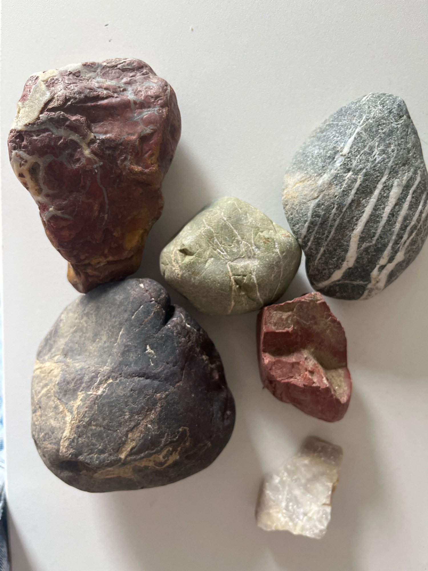 A collection of seven rocks are arranged on a white surface, likely a table or countertop, in a top-down view. The rocks vary in size, shape, color, and texture.
Starting from the top left and moving clockwise:
1. A large, irregularly shaped rock with a reddish-brown base and swirling patterns of white and lighter red. It appears to be a rough-textured stone.
2. A medium-sized, oval-shaped gray rock with distinct white parallel stripes running diagonally across its surface. The texture appears smoother than the first rock.
3. A small, greenish-gray, rounded rock with faint lighter green veining.
4. A small, irregularly shaped reddish-brown rock with a rough, broken surface.
5. A very small, translucent, irregularly shaped white or clear crystal-like rock.
6. A large, dark gray to black, rounded rock with subtle lighter gray and tan veining. The texture seems somewhat rough and uneven.
7. A medium-sized, pale green, rounded rock with subtle veining.
The lighting suggests a natural light source from above and to the left, casting subtle shadows. The background is a plain white surface.
The image does not provide any information or context about the city of Kraljevo, Serbia, or any specific location within it. There are no people, actions, or interactions depicted. The scene is purely a still-life arrangement of geological specimens.