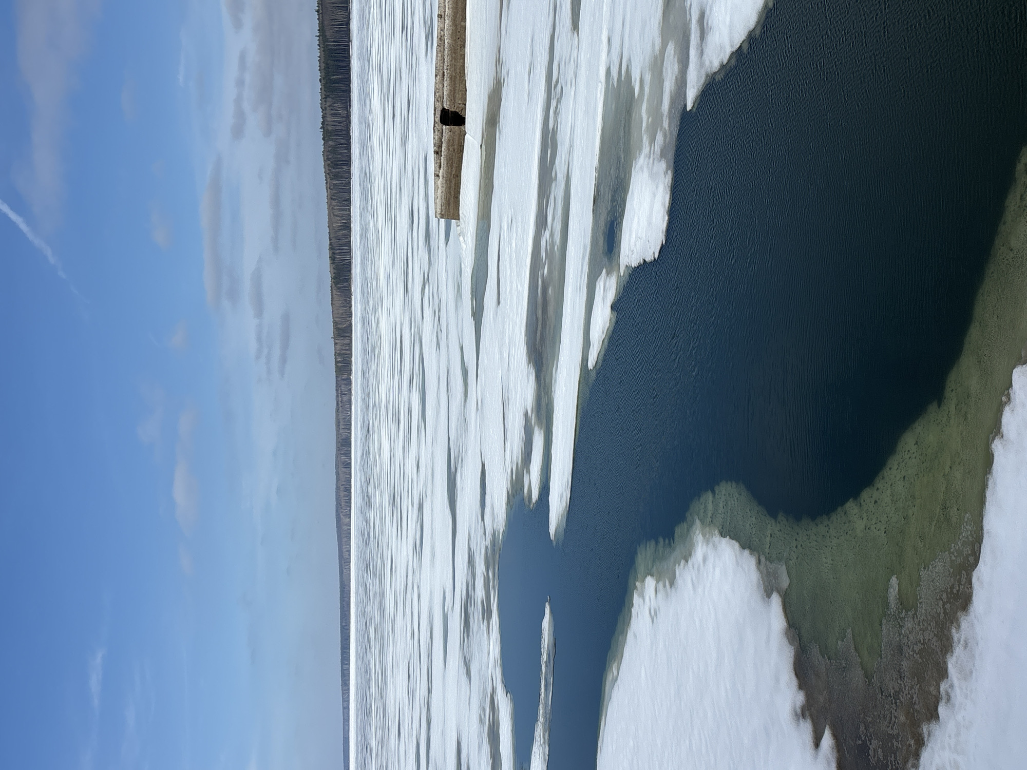 The image shows a partially frozen body of water, likely a lake or river, in Chaikovsky, Russia. Patches of open water are visible amidst large expanses of ice and snow cover. A shoreline with a dense line of trees stretches along the horizon. A wooden structure, possibly a pier or dock, is partially covered in ice and snow. The sky is partly cloudy with a prominent contrail.